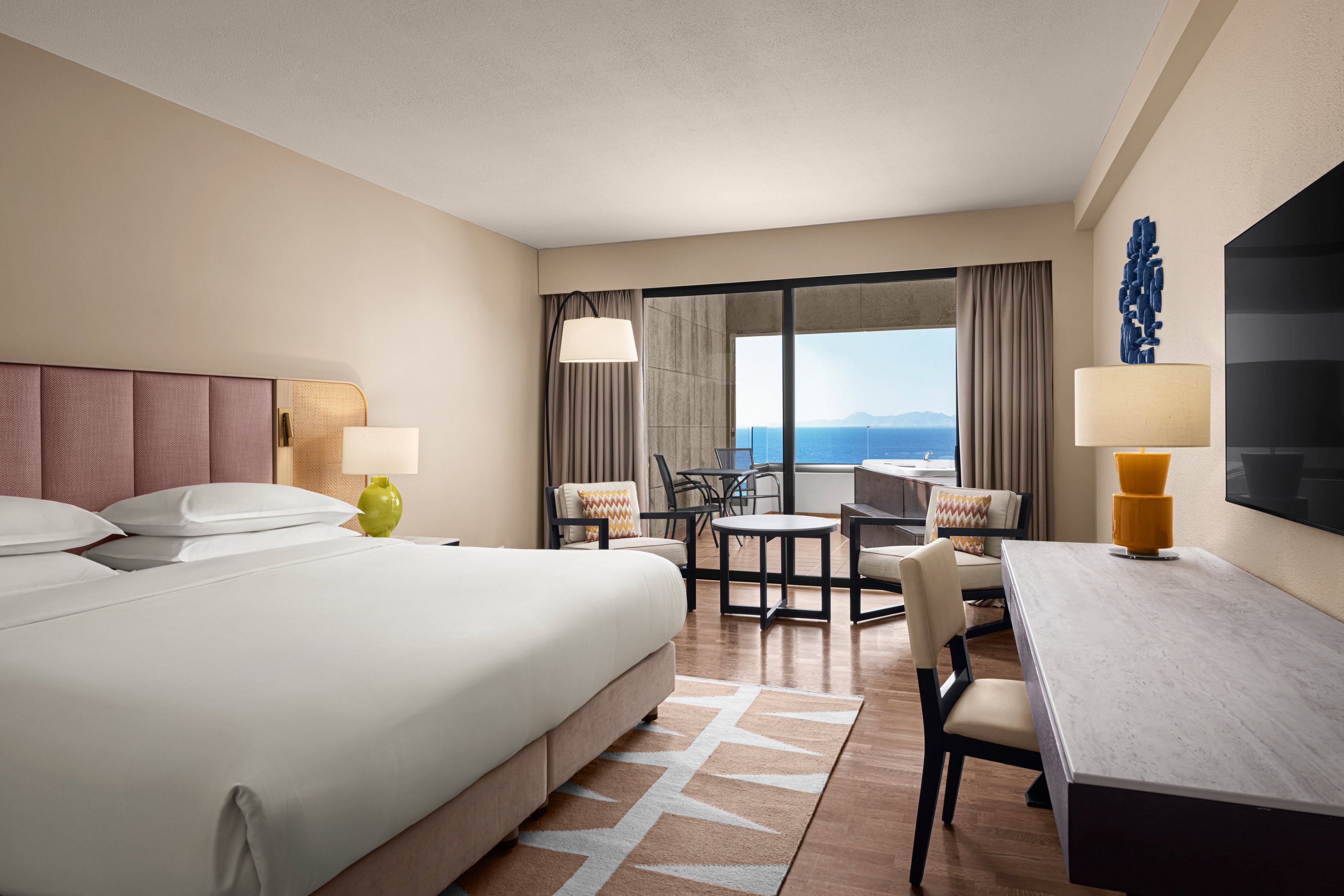 A contemporary hotel room features a large bed with crisp white linens and upholstered headboard. A desk, chairs, and side tables with lamps create a comfortable workspace and seating area. Floor-to-ceiling glass doors open to a balcony with chairs and a table overlooking a bright blue ocean and distant coastline. Neutral walls, warm wood flooring, and soft accent colors give the space a calm, inviting atmosphere.