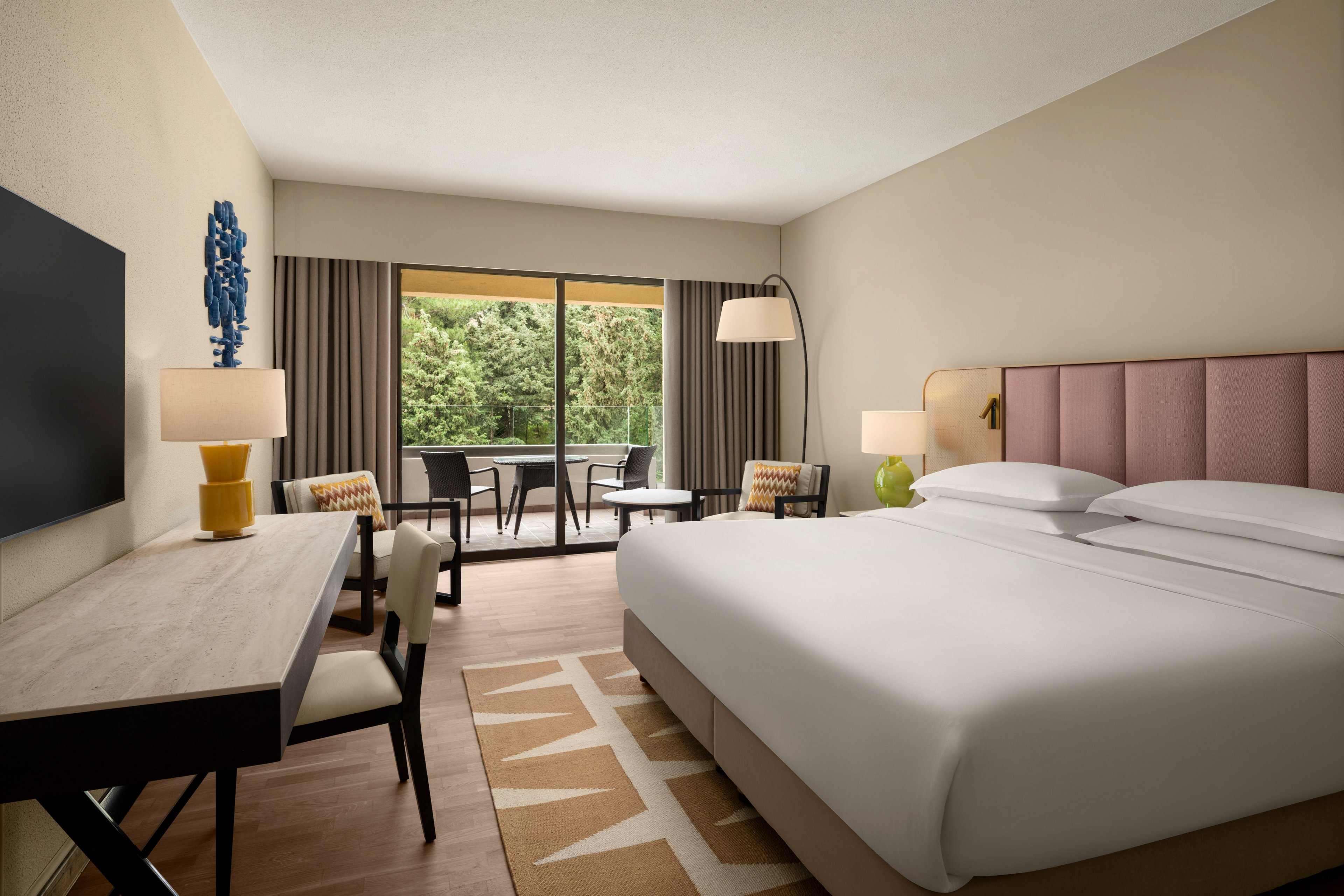 A contemporary hotel room features a large bed with white linens and a padded headboard. A desk with a flat-screen television and a floor lamp sit opposite the bed, while two accent chairs flank a small table near the window. Sliding glass doors open to a balcony with outdoor seating overlooking lush greenery. Neutral walls, patterned rug, and coordinated decor create a calm, inviting atmosphere.