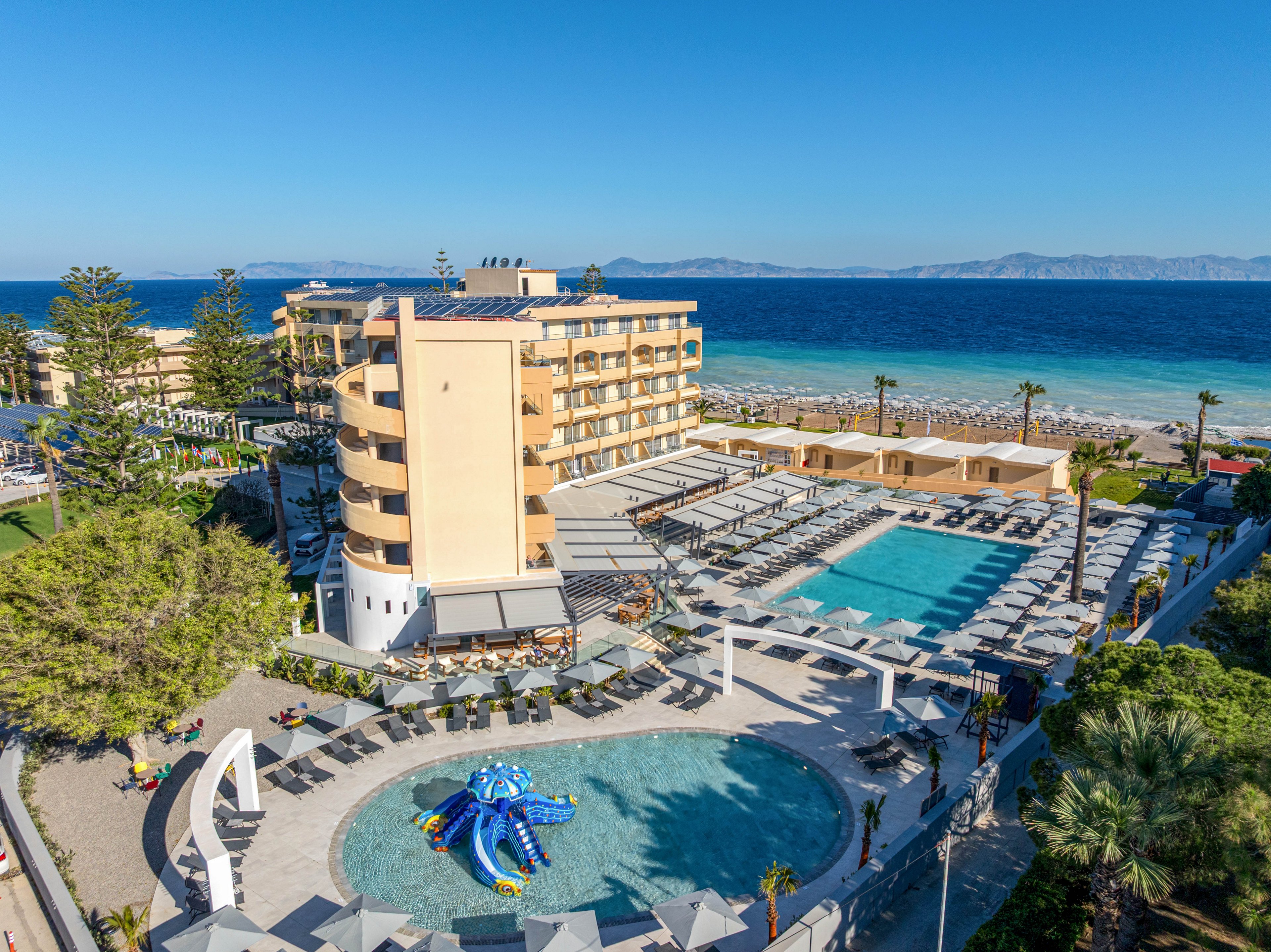 A large seaside resort complex features multiple outdoor swimming pools and sun loungers arranged in neat rows. The main building rises several stories high beside the coastline, offering views of the blue sea and distant mountains. A circular children’s pool with a colorful waterslide structure sits in the foreground, surrounded by shaded seating areas. Clear skies and bright sunlight create a relaxed vacation atmosphere at this beachfront destination.
