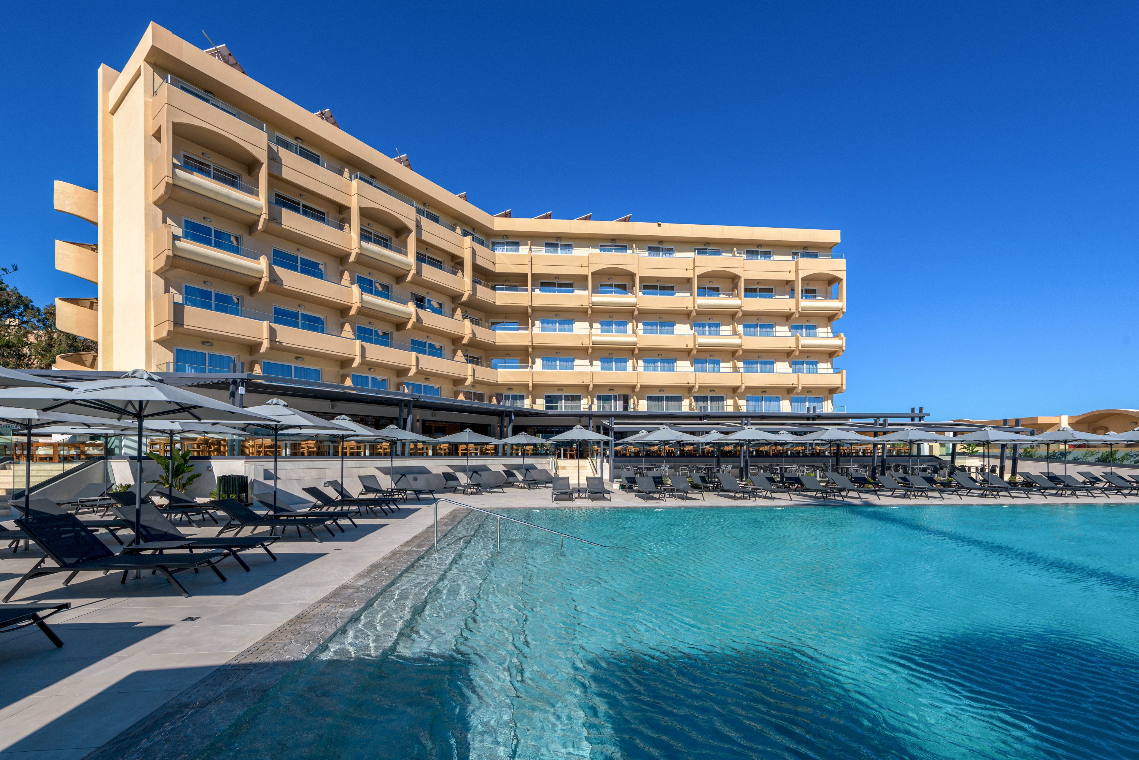 A contemporary multi-story hotel stands beside a large outdoor swimming pool under a clear blue sky. Rows of sun loungers and umbrellas line the poolside area, suggesting a resort setting for relaxation. The building features multiple balconies facing the water, offering guests views of the pool and surrounding landscape. Bright sunlight and the turquoise color of the pool create a calm, vacation-like atmosphere.