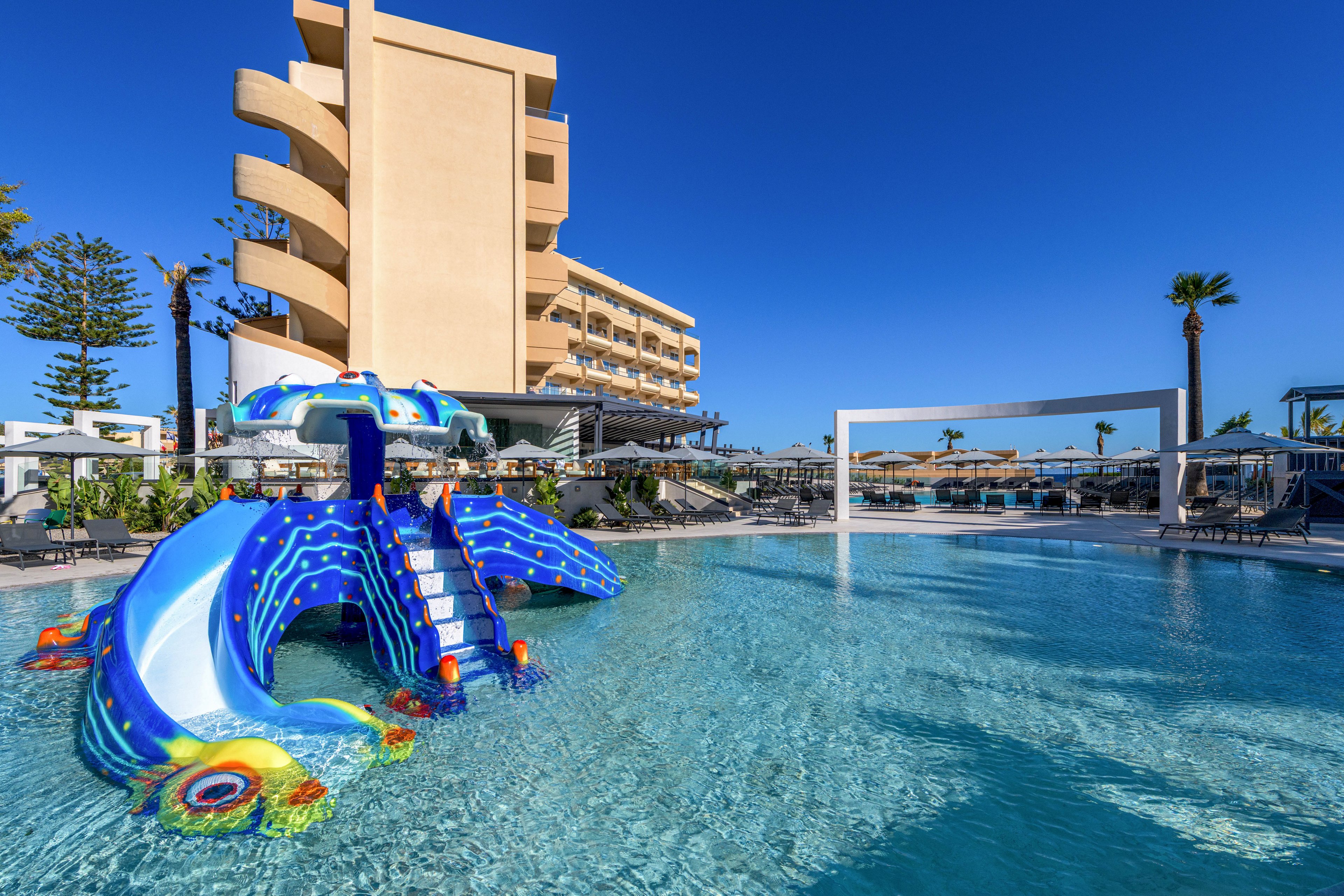 A bright blue dragon-shaped waterslide stands in the shallow area of a large resort swimming pool. The pool is surrounded by sun loungers, umbrellas, and modern outdoor structures under a clear blue sky. A multi-story hotel building rises in the background, with balconies overlooking the pool area. The scene suggests a family-friendly vacation setting with playful water attractions and sunny weather.