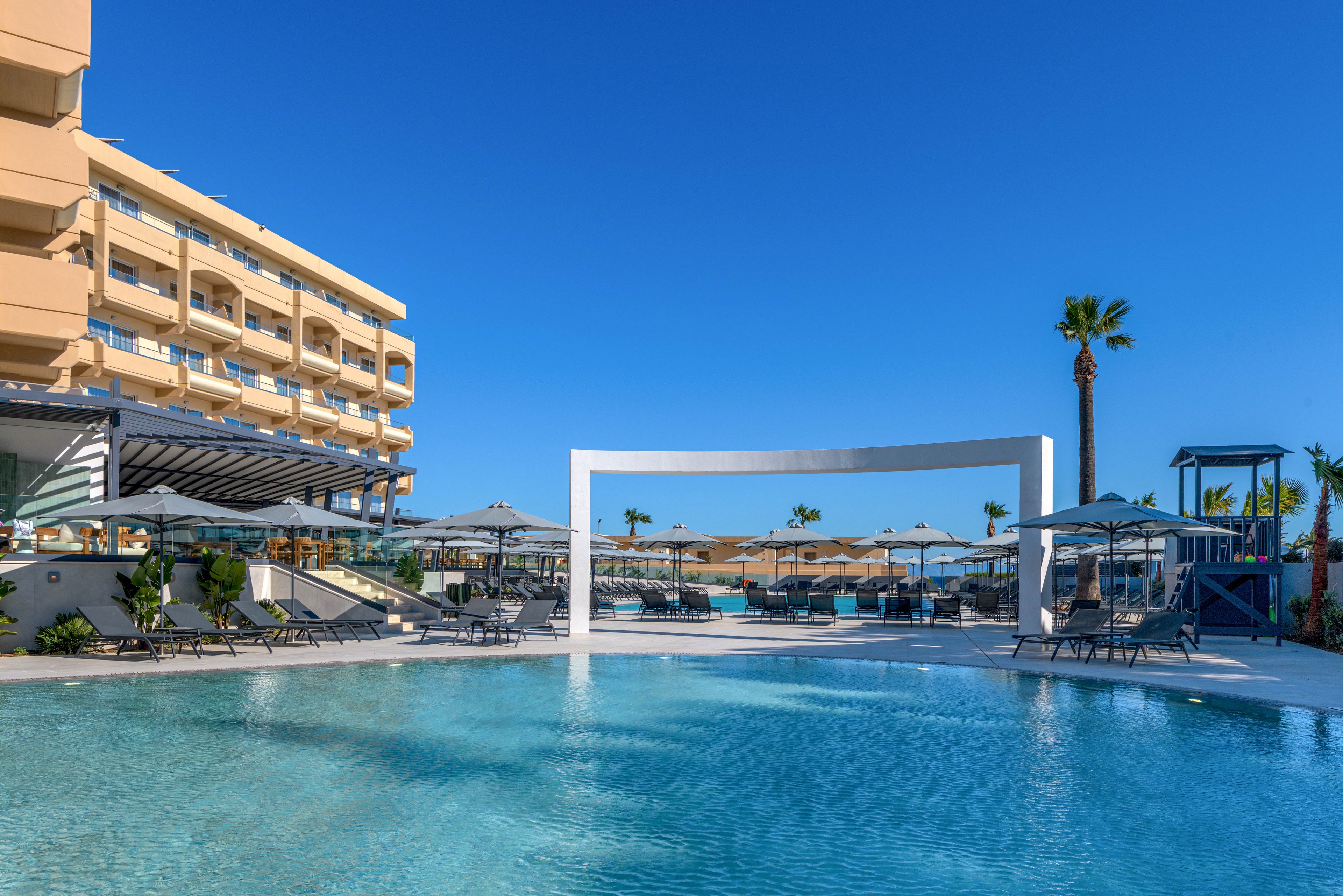 A contemporary resort features a large, inviting swimming pool in the foreground. Sun loungers and umbrellas line the poolside, creating a relaxing vacation atmosphere. A multi-story hotel building stands to the left, while a white rectangular frame structure adds a modern architectural accent near the water. The bright blue sky and palm trees emphasize the warm, sunny climate and leisure setting.