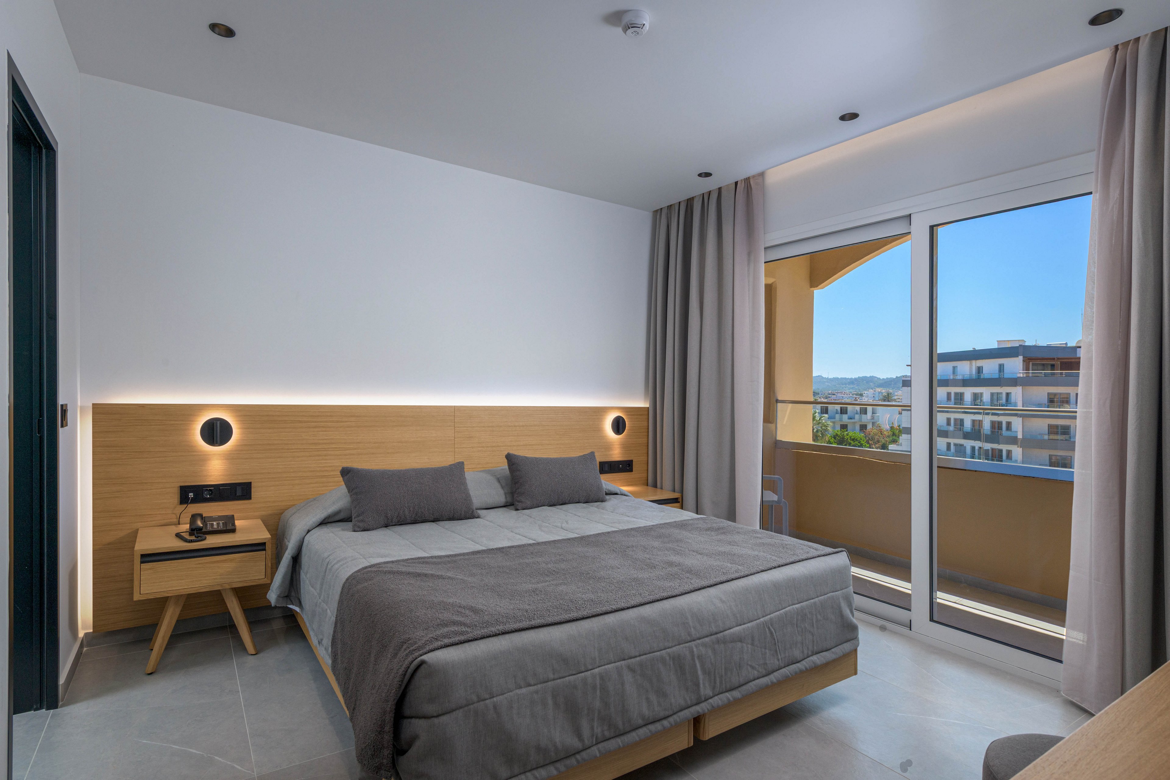 A contemporary hotel bedroom features a large double bed with gray bedding and matching pillows. Wooden nightstands with lamps and a digital alarm clock flank the bed, set against a minimalist headboard with integrated lighting. Floor-to-ceiling curtains frame a sliding glass door that opens to a balcony overlooking neighboring buildings and a distant landscape. Recessed ceiling lights and neutral tones create a calm, modern atmosphere.