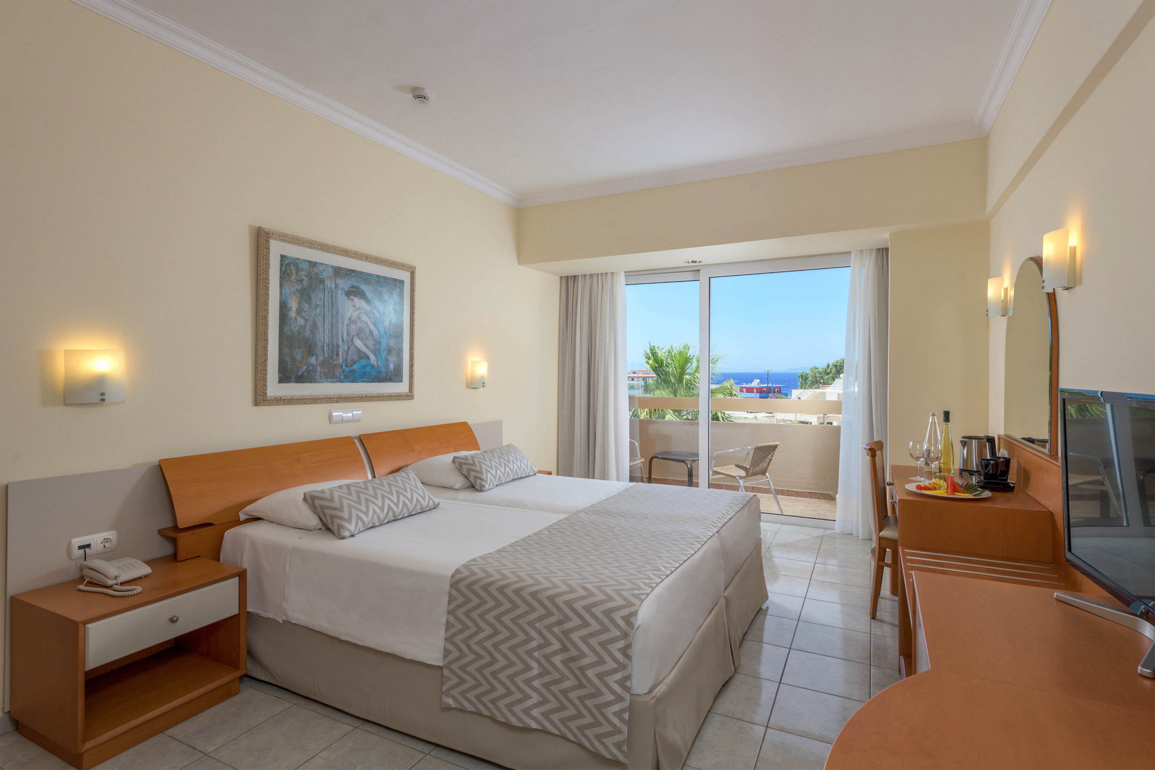 A neatly made double bed with patterned runners is the focal point of this bright hotel bedroom. The room features light-colored walls, tiled flooring, and simple wooden furniture including bedside tables, a desk, and a chair. Large glass doors open onto a balcony with outdoor seating and a partial view of the sea and greenery. Soft wall lighting and framed artwork above the bed contribute to a calm, inviting atmosphere.