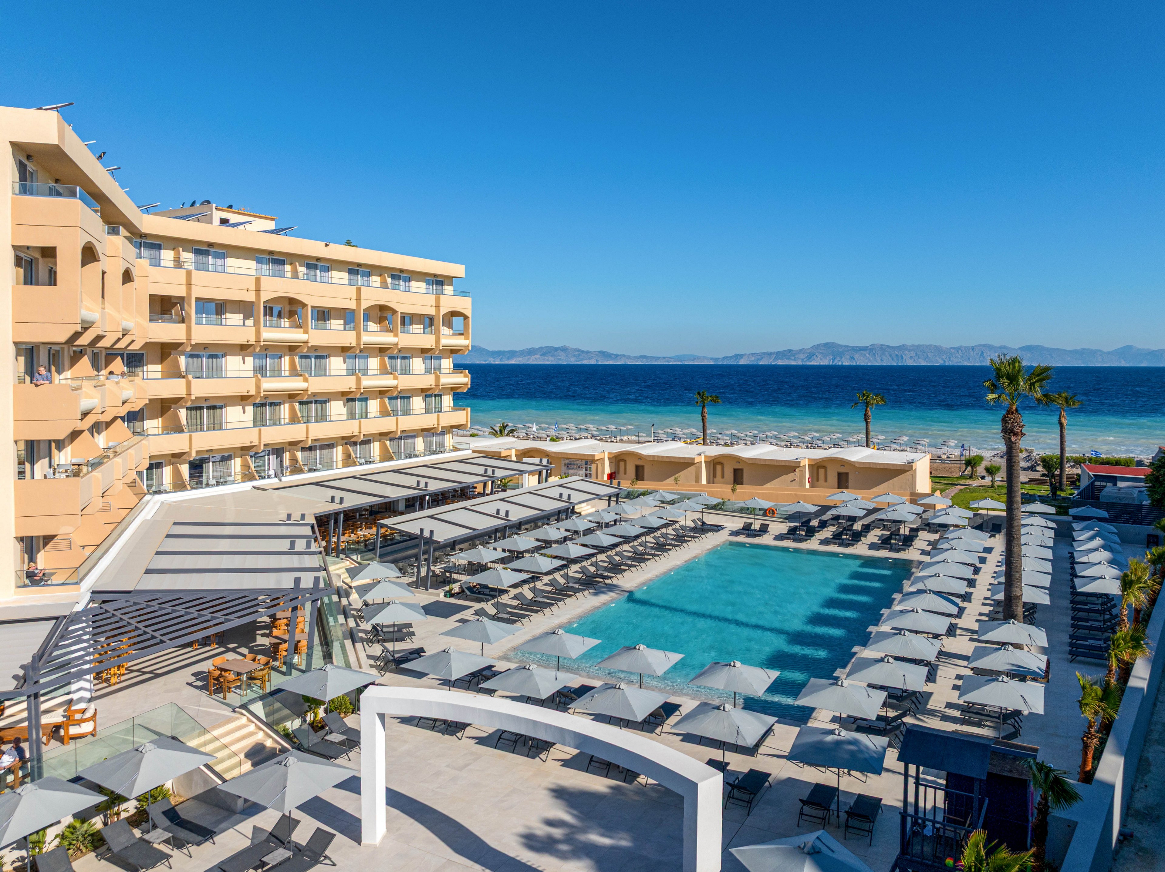 A modern seaside resort hotel overlooks a large rectangular swimming pool filled with clear blue water. Numerous sun loungers and umbrellas are neatly arranged around the poolside terraces, creating an organized vacation setting. The multi-story beige building faces the ocean, with balconies offering views of the coastline and distant mountains. Bright sunlight and a cloudless blue sky emphasize a relaxed, summery atmosphere by the sea.