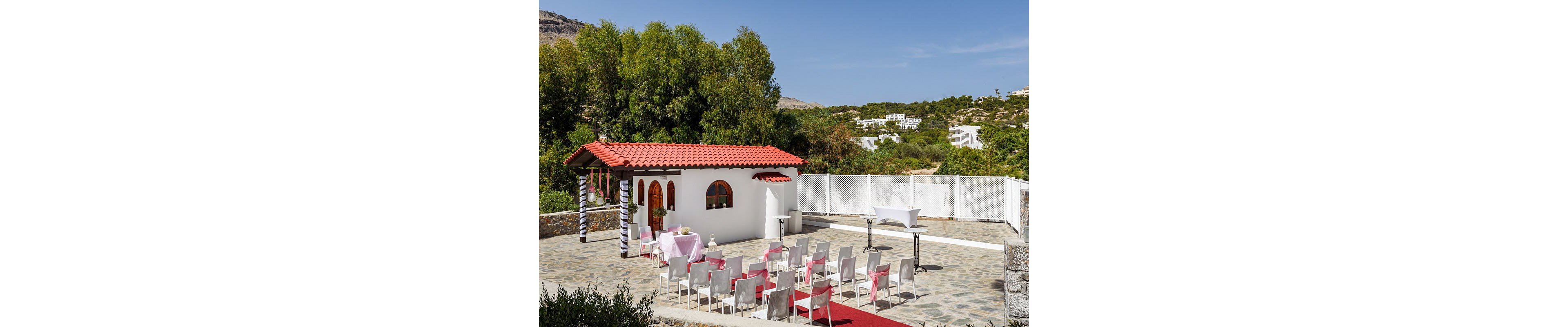 An intimate outdoor wedding ceremony is arranged in front of a small white chapel with a red tiled roof. Rows of white chairs with pink sashes line a stone courtyard, facing a simple altar table and backdrop. A red carpet aisle leads toward the chapel entrance, framed by striped columns and arched wooden doors and windows. Lush green trees and distant white buildings on the hillside complete the sunny, Mediterranean-style setting.