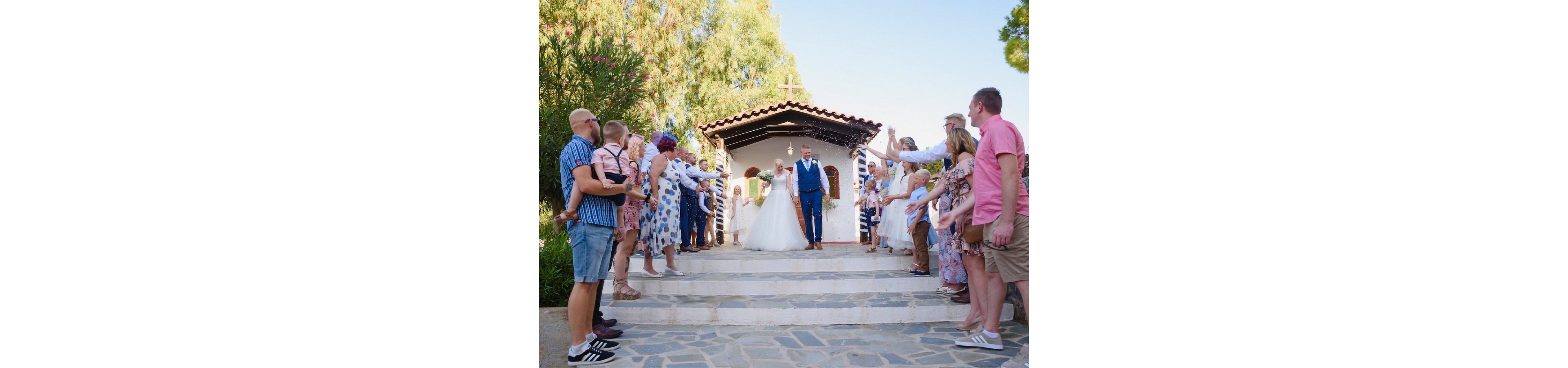 A bride and groom walk down the steps of a small white chapel as friends and family stand on either side. The guests form a celebratory corridor, raising their arms and tossing confetti toward the couple. The scene takes place outdoors in bright daylight, with trees and stone paving surrounding the chapel. The mood is joyful and festive, capturing a traditional wedding moment.