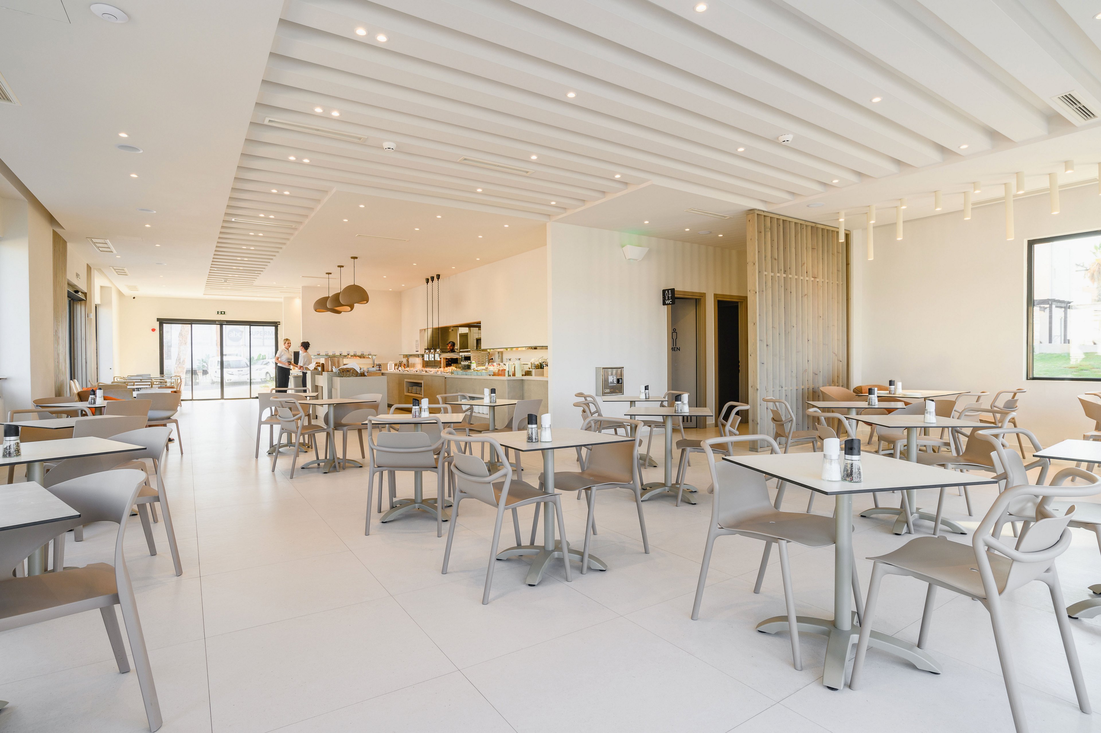 Spacious contemporary restaurant interior with neatly arranged tables and chairs. The room is brightly lit with large windows and recessed ceiling lights. Neutral tones and minimalist decor create a clean, airy atmosphere. A counter area and a few staff members are visible in the background, suggesting a ready but currently unoccupied dining space.