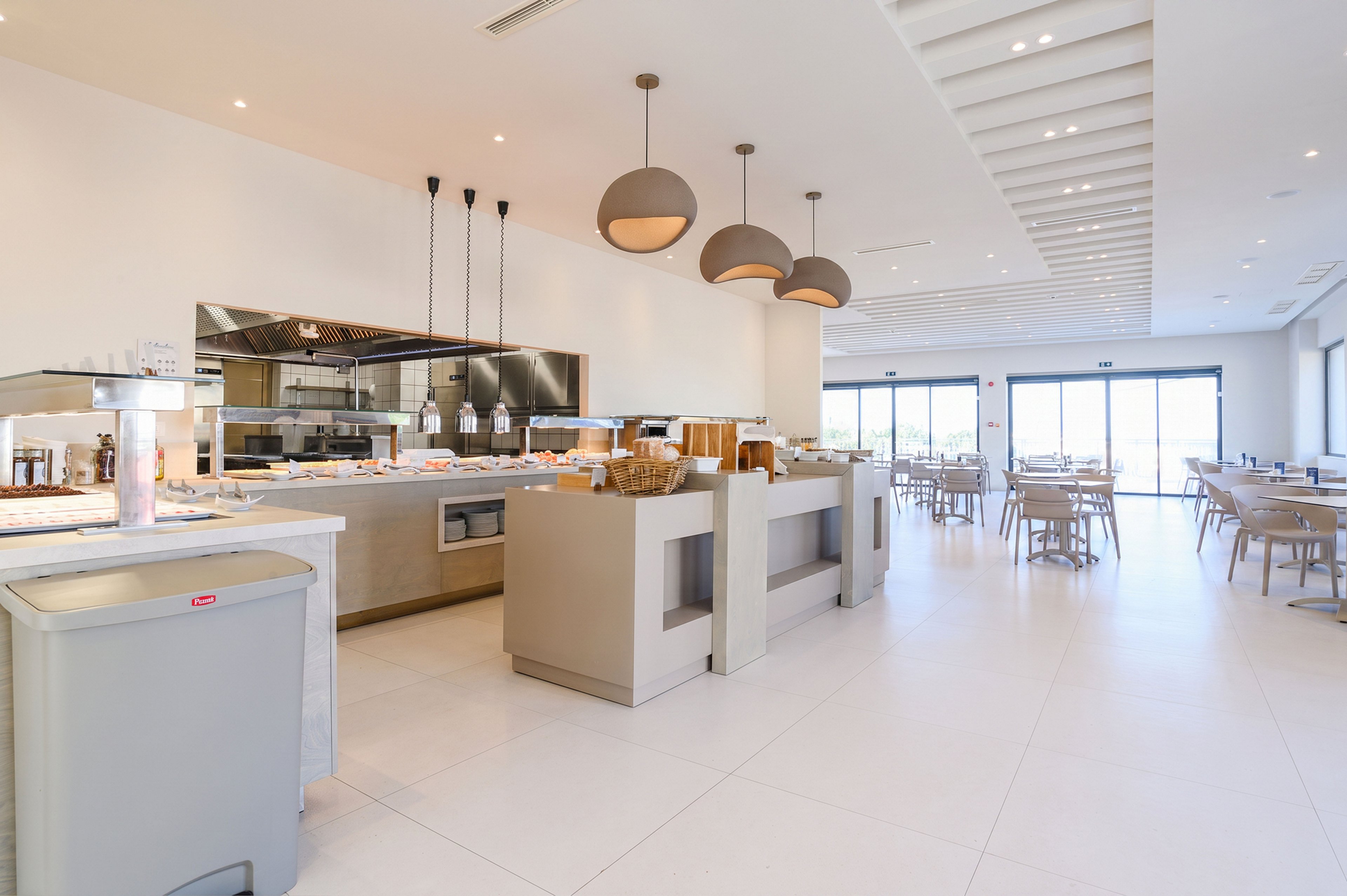 Spacious contemporary restaurant interior with a central self-service counter. Open kitchen equipment and food stations are visible behind the counter area. Large windows at the back let in natural light, illuminating the dining tables and chairs. Neutral colors, clean lines, and minimalist decor create a bright, modern atmosphere.