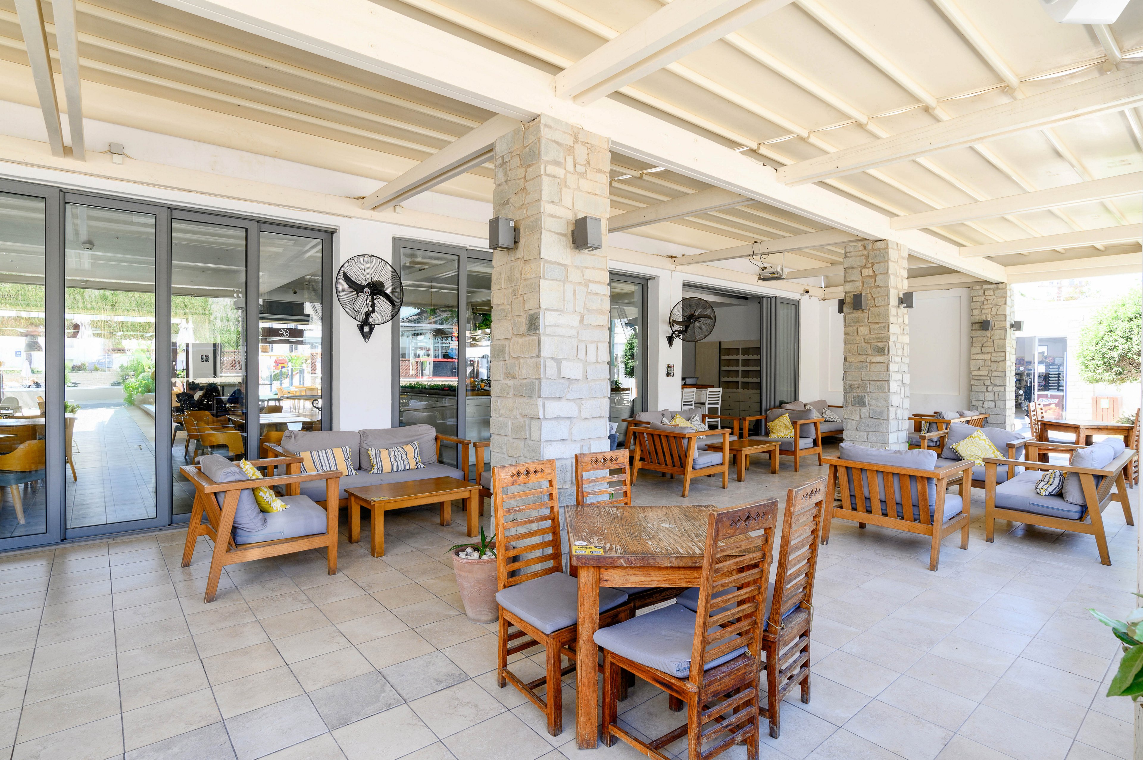 A bright, modern outdoor patio features wooden tables and cushioned lounge seating arranged under a covered roof. Large glass doors line one side, connecting the terrace to an indoor area. Stone pillars, ceiling fans, and neutral-toned tiles create a relaxed, contemporary atmosphere. The space appears ready for guests, with neatly arranged chairs and decorative plants adding warmth.