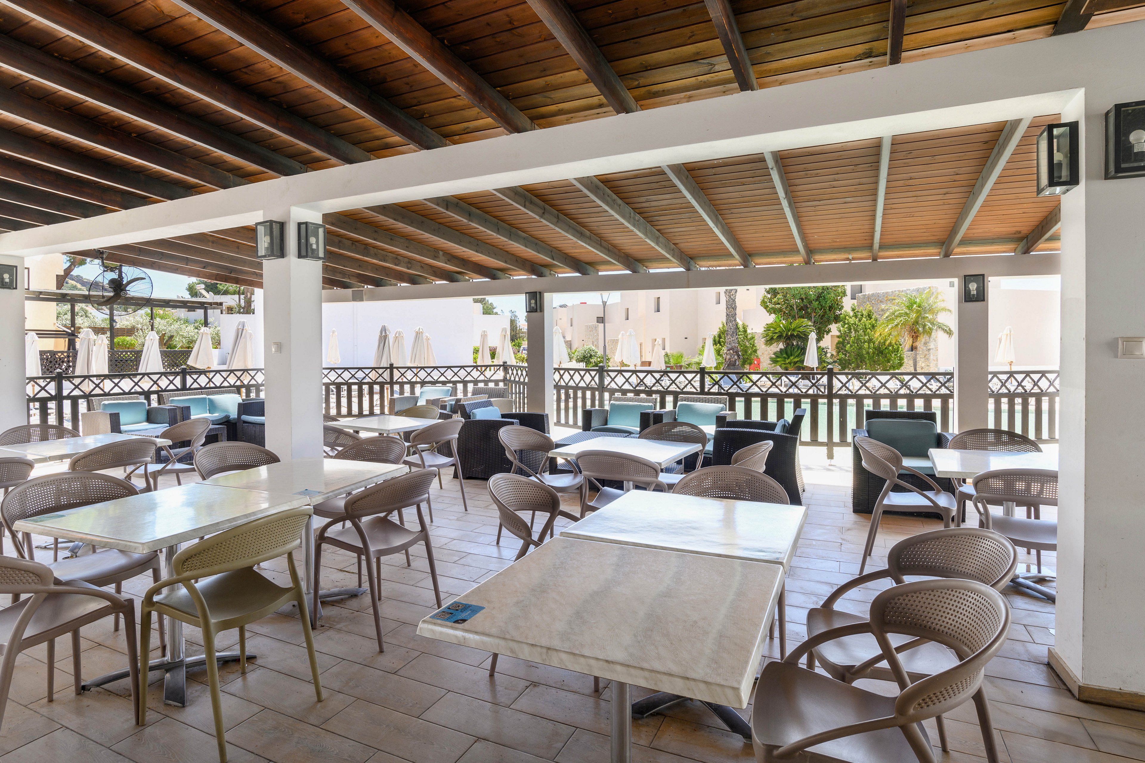 An empty outdoor cafe or restaurant seating area is arranged under a wooden roof. Light-colored tables and modern plastic chairs are neatly organized in rows on a tiled floor. The space opens toward a bright courtyard with greenery, white buildings, and sun umbrellas in the background. Natural daylight filters through the open sides, creating a calm and relaxed atmosphere.