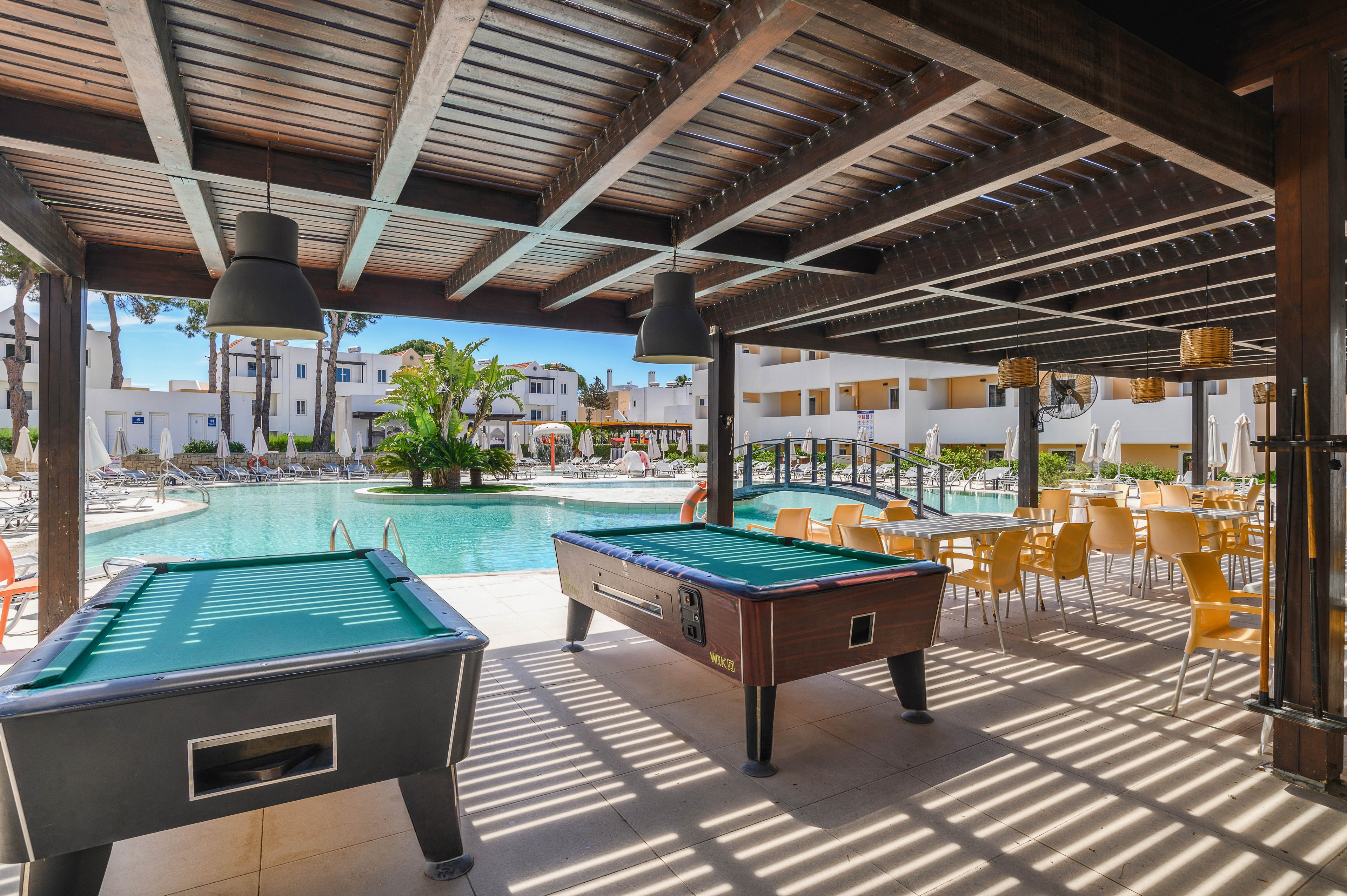 An open-air lounge area features two billiard tables under a wooden pergola. Beyond the tables, a resort-style swimming pool is surrounded by sun loungers and white buildings. A small bridge crosses part of the pool, adding visual interest to the scene. To the right, rows of yellow chairs and tables create a casual outdoor dining space, all bathed in bright daylight.