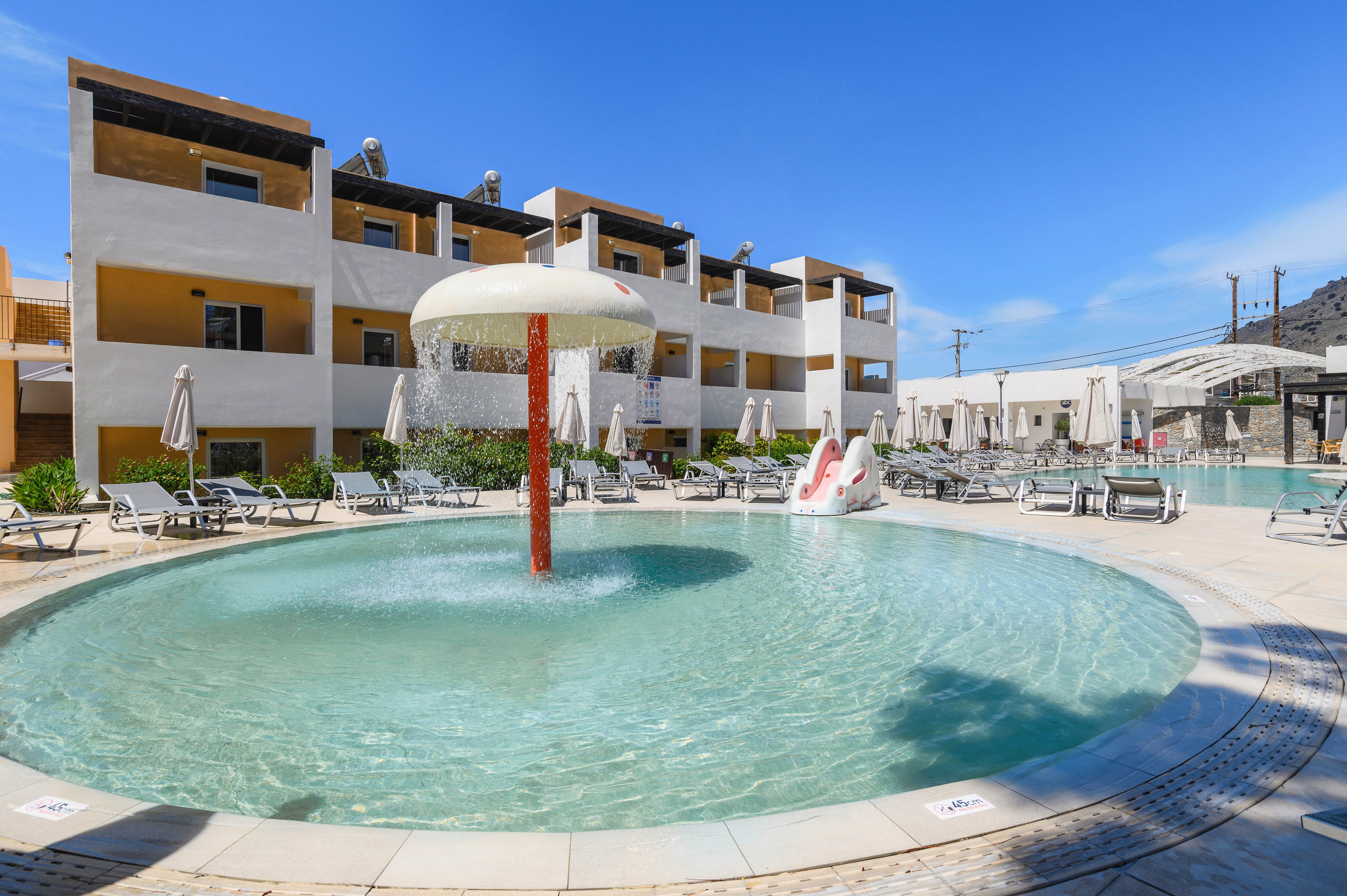 A shallow circular kiddie pool features a central mushroom-shaped fountain spraying water. The pool is surrounded by sun loungers and umbrellas in a modern resort courtyard. In the background, a small water slide shaped like a white animal adds a playful element. Multi-story beige and white buildings frame the pool area under a clear blue sky, creating a bright and inviting vacation setting.