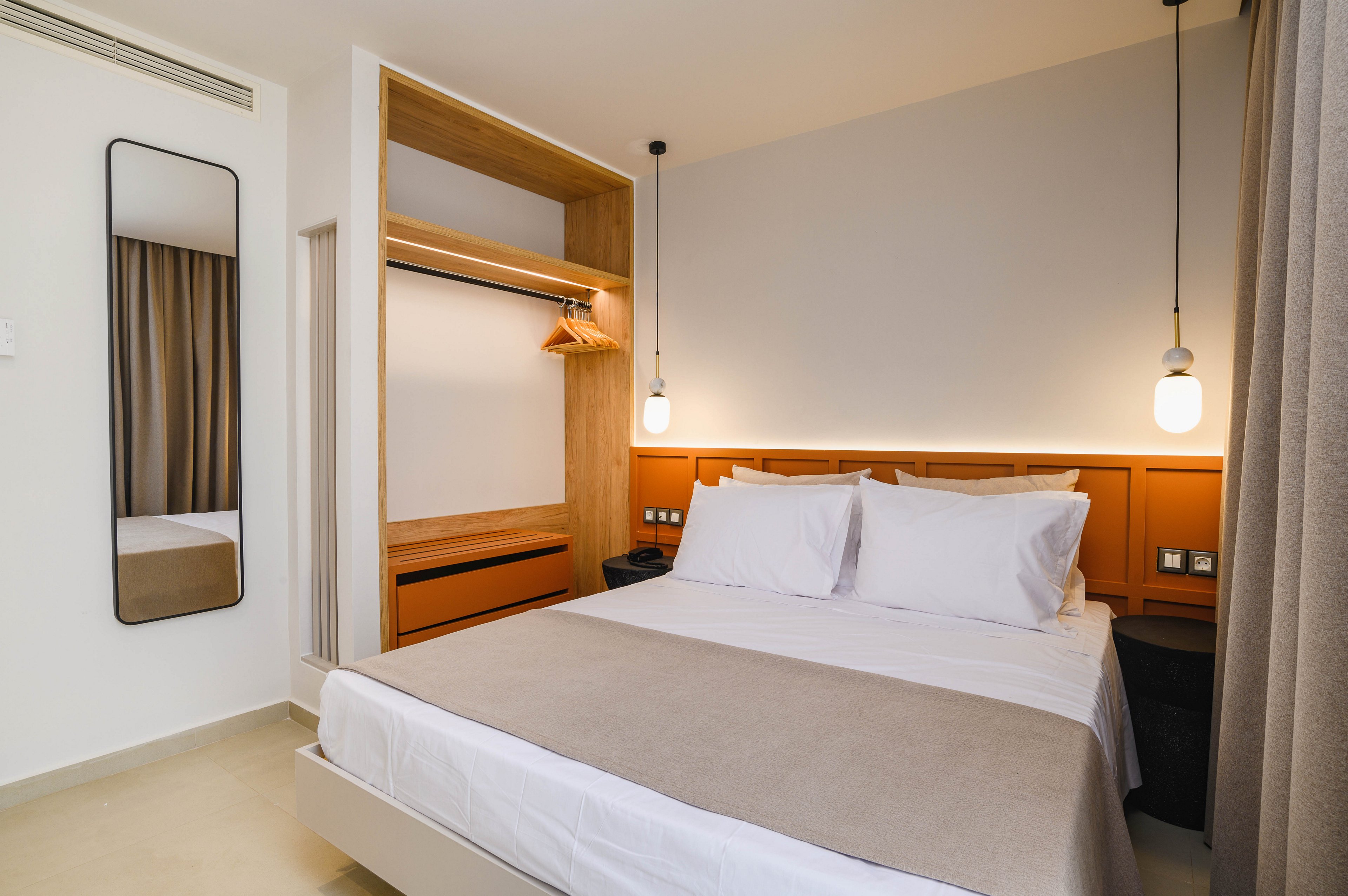 A neatly made double bed with white linens and a beige throw is the focal point of this modern hotel bedroom. The room features a built-in open wardrobe with wooden shelves and a hanging rail, along with a single wooden hanger. Warm lighting from two pendant lamps and a concealed headboard light creates a cozy atmosphere. A tall wall mirror and floor-to-ceiling curtains add to the clean, minimalist design.