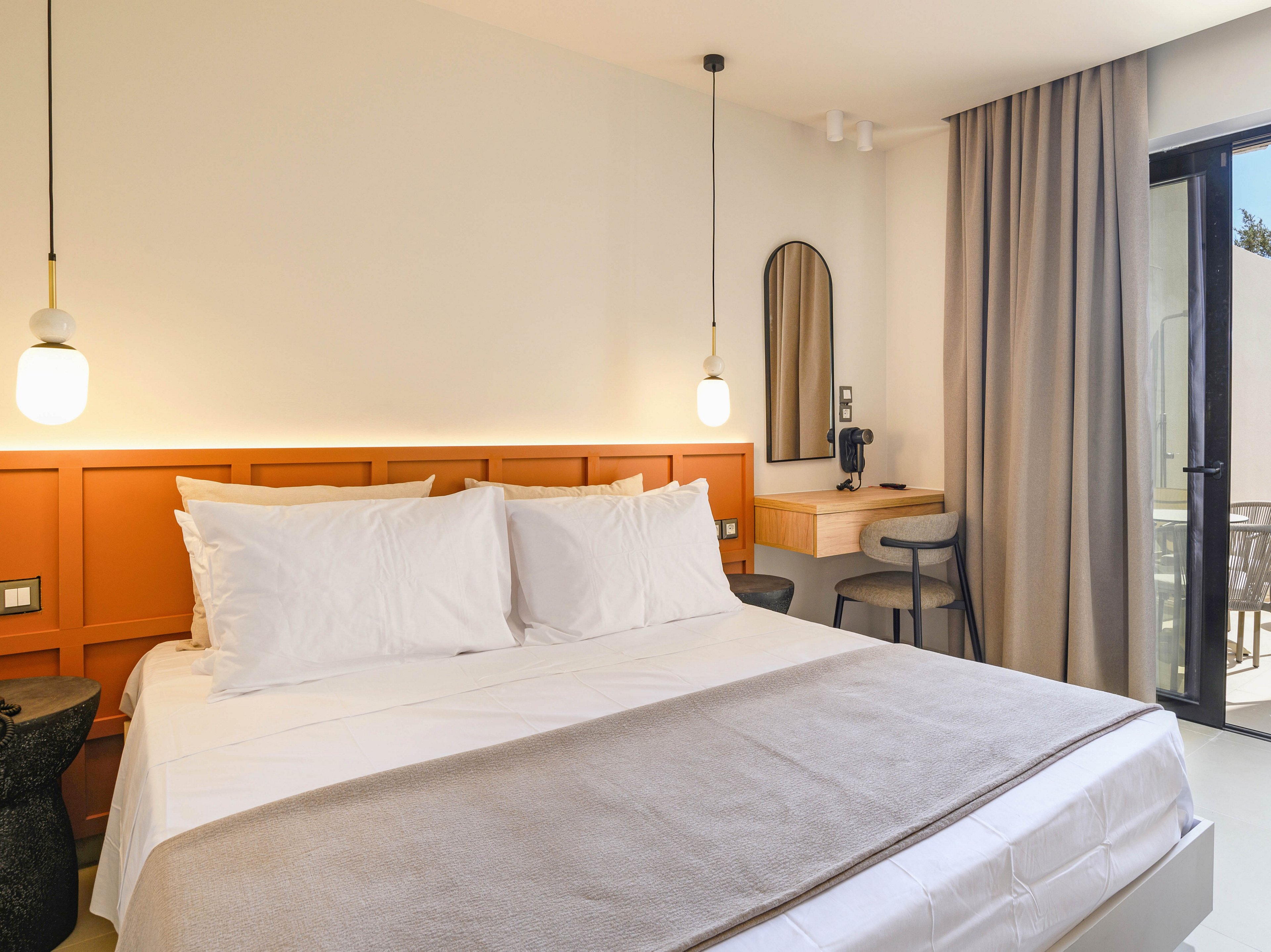 A contemporary hotel bedroom features a neatly made double bed with crisp white linens and a light beige throw. Warm orange paneling forms a headboard accent wall, complemented by two hanging pendant lights. A small wooden desk with a mirror and chair sits in the corner, creating a compact workspace or vanity area. Floor-to-ceiling curtains frame a glass door that opens to an outdoor balcony, allowing natural light to brighten the neutral interior.