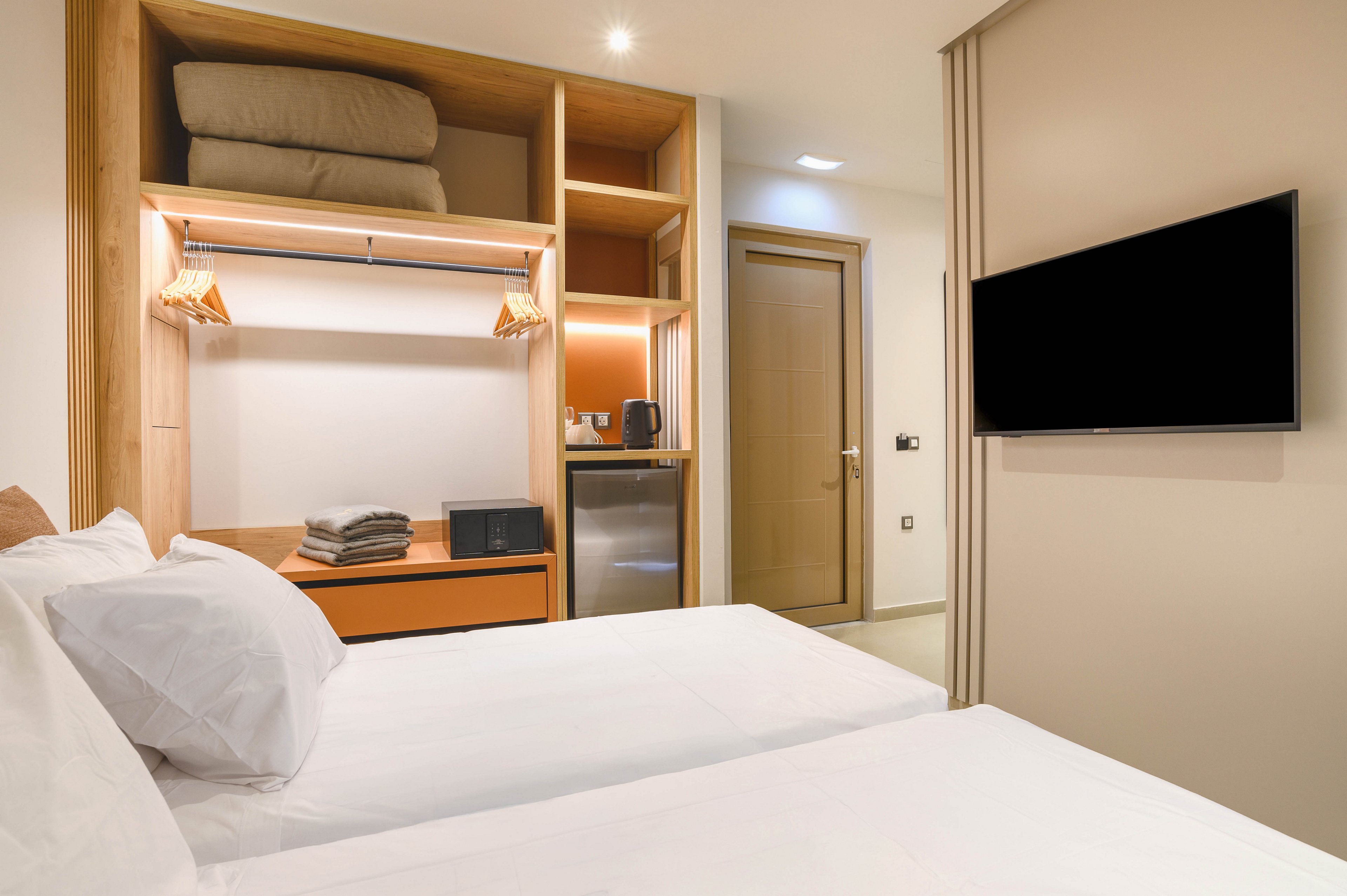 A contemporary hotel room features two neatly made twin beds in the foreground. An open wardrobe with shelves, folded bedding, hangers, and stacked towels is built into the wall. A compact minibar area with a small refrigerator, kettle, and cups sits beneath warm accent lighting. A flat-screen television is mounted on the opposite wall, and a closed door leads to an adjoining space, creating a clean and minimalist atmosphere.