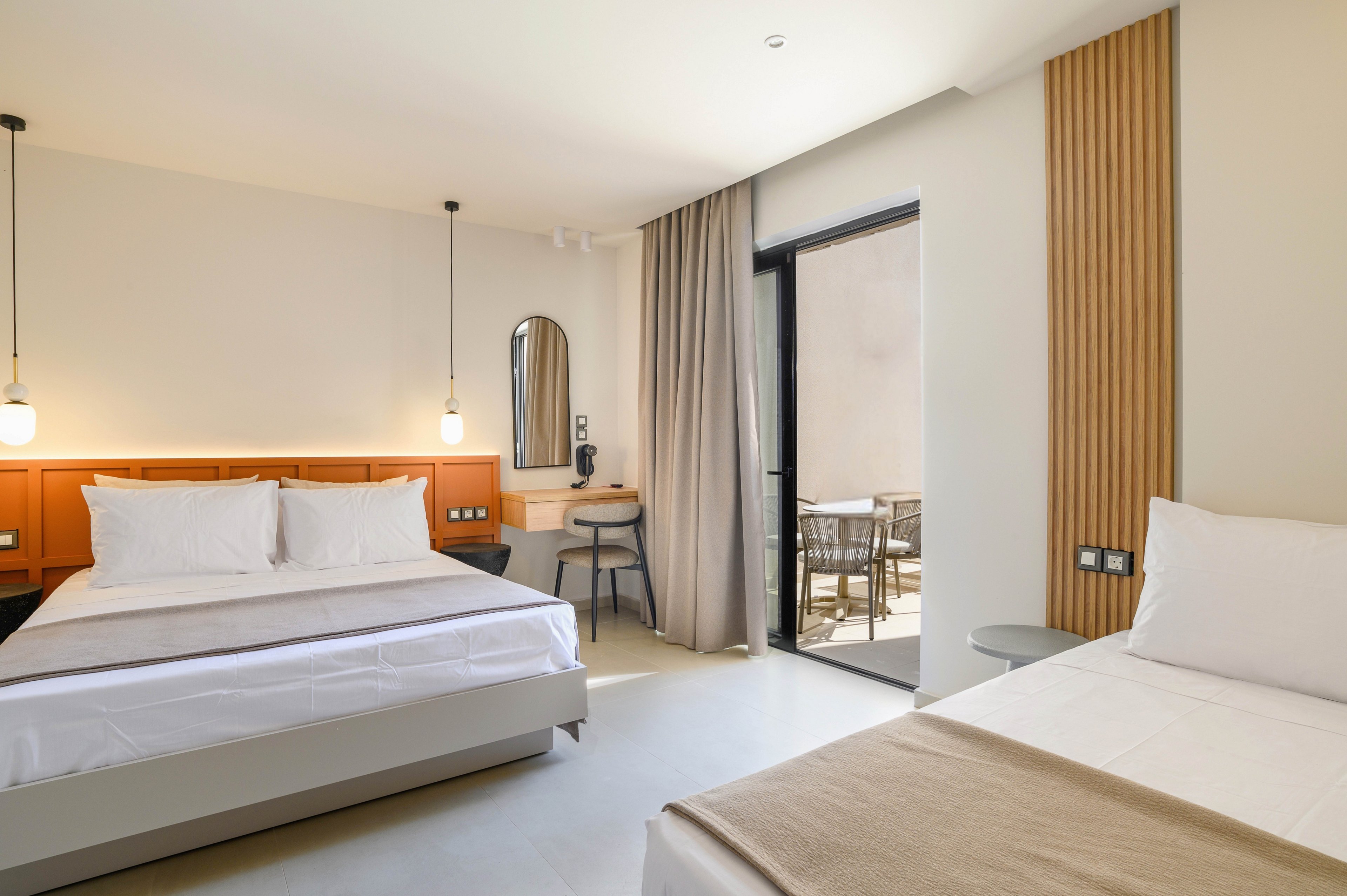 A contemporary hotel room features two neatly made beds with white linens and beige throws. The space includes a wooden headboard, pendant lights, and a small desk with a mirror. Floor-to-ceiling curtains frame a glass door that opens to a private balcony with outdoor seating. Neutral tones and clean lines create a calm, minimalist atmosphere.