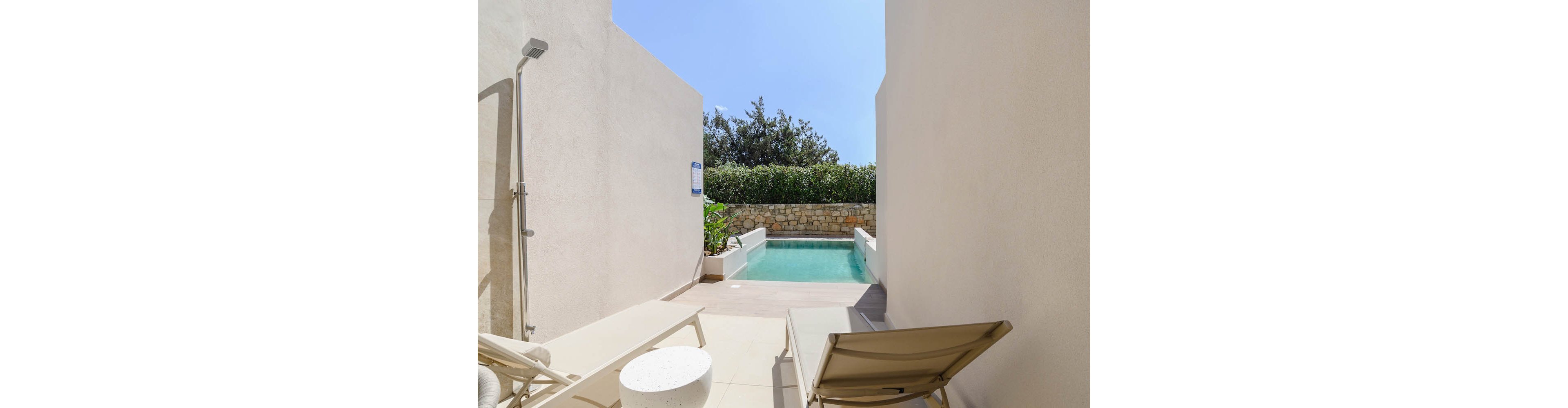 A small private plunge pool is nestled between two light-colored walls in a modern courtyard. Two sun loungers and a round side table sit on the tiled patio, facing the clear blue water. An outdoor shower is mounted on the left wall, adding to the resort-like feel. Green shrubs and a stone wall border the far end of the pool under a bright blue sky.