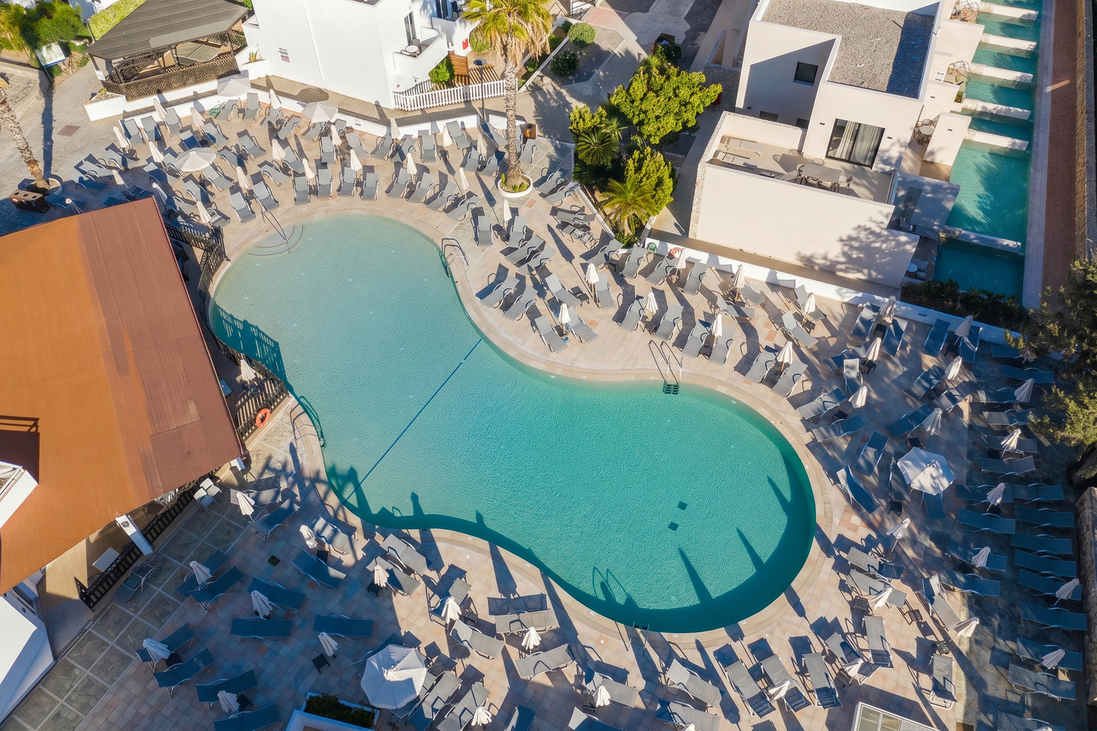An aerial view shows a large, curving swimming pool at a modern resort. Numerous empty lounge chairs and umbrellas surround the pool in neat rows, creating a symmetrical pattern. The surrounding buildings are white and contemporary, with palm trees adding a touch of greenery. Bright daylight and clear shadows suggest a sunny, warm climate and a tranquil, off-peak moment.