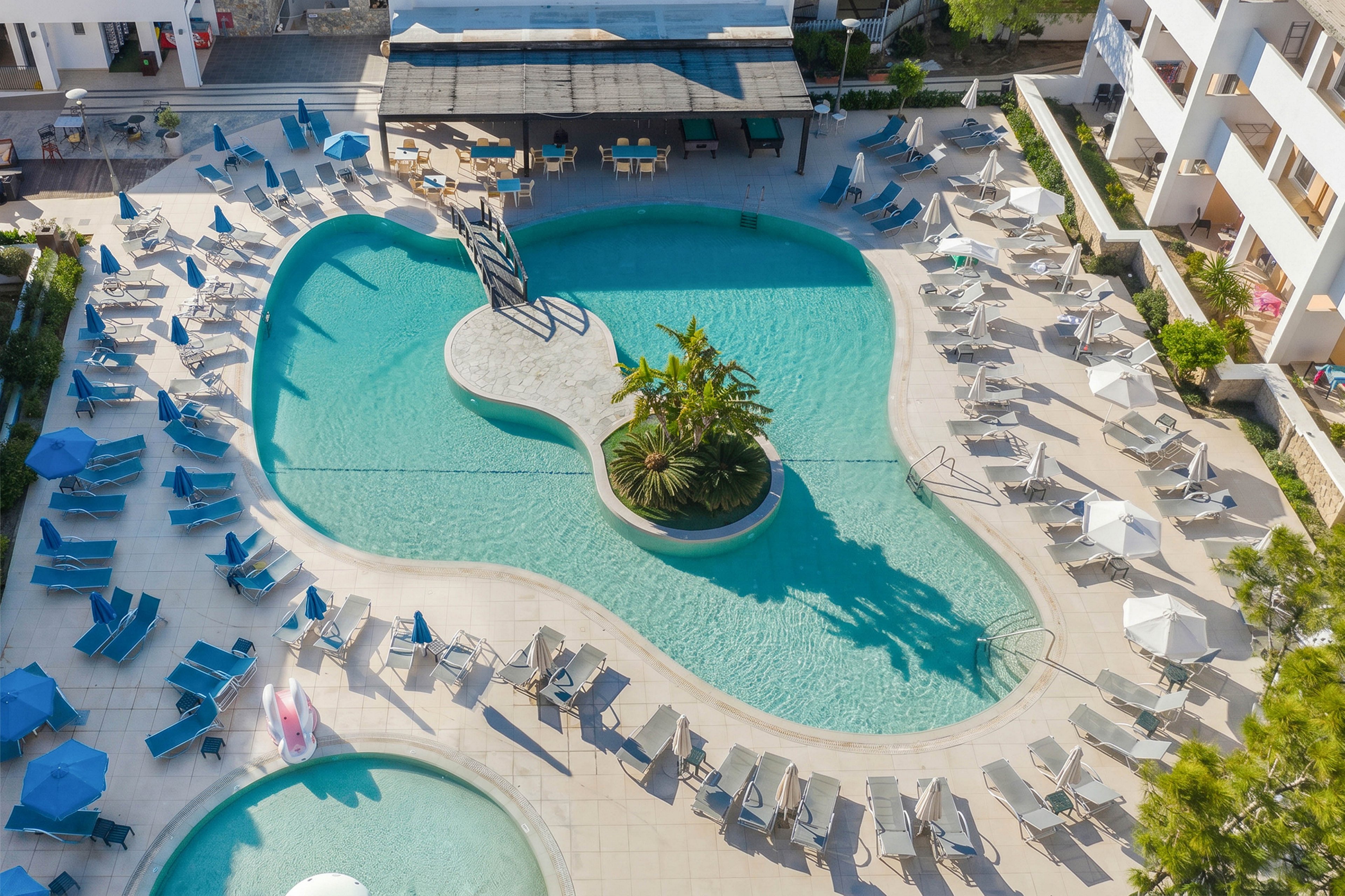 An aerial view shows a large resort swimming pool with a curved, organic shape and a central island of tropical plants. Numerous lounge chairs and umbrellas surround the pool, creating a structured pattern on the pool deck. A smaller circular pool is visible in the foreground, suggesting a separate relaxation or children’s area. Modern white buildings and a shaded bar or seating area frame the scene, evoking a sunny vacation atmosphere.