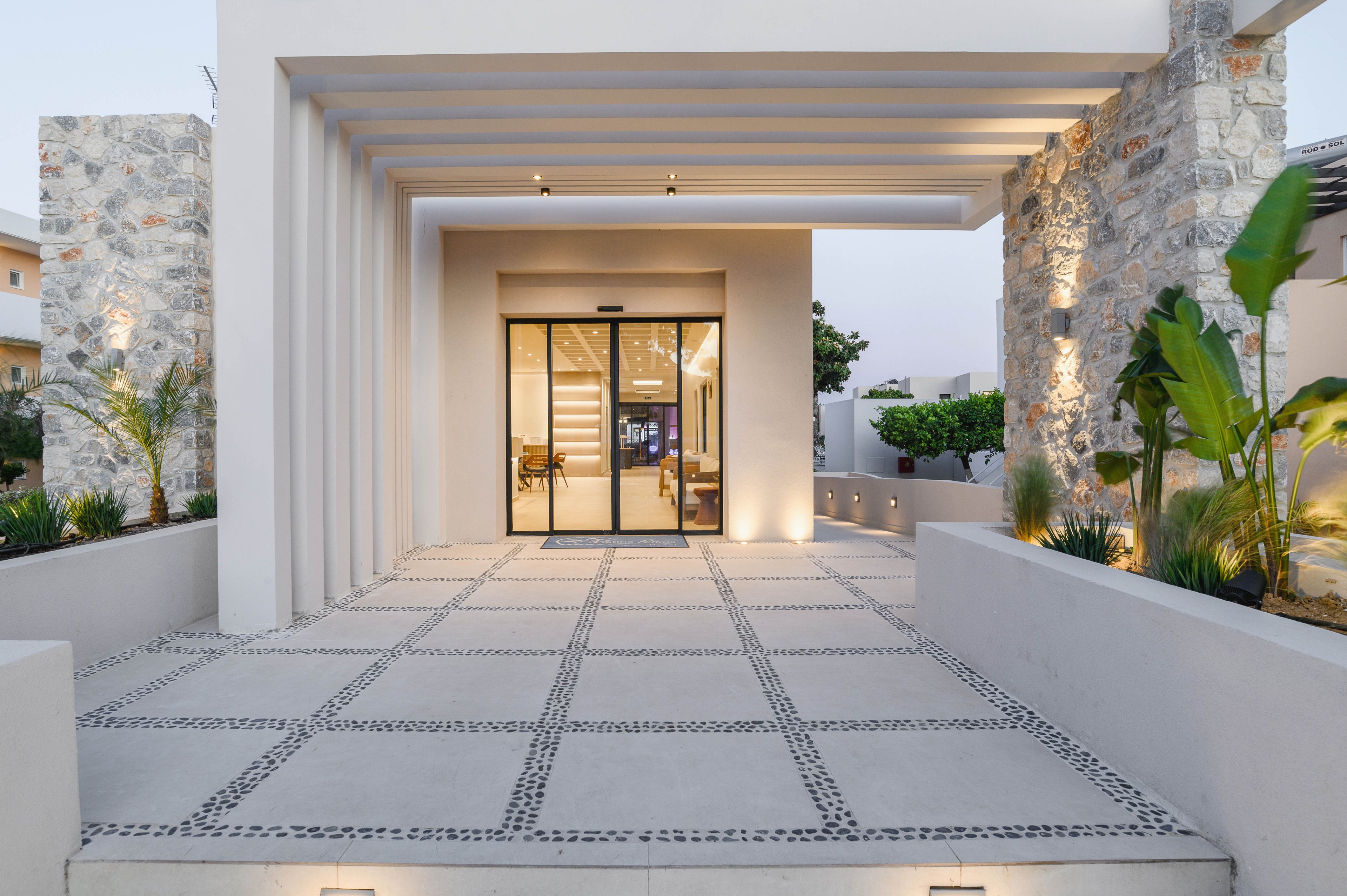 A contemporary building entrance features a wide tiled walkway leading to glass double doors. Linear beams create a shaded portico, casting subtle shadows across the minimalist facade. Stone walls and integrated planters with greenery add texture and warmth to the clean architectural lines. Soft exterior lighting highlights the pathway and structural details in the early evening light.