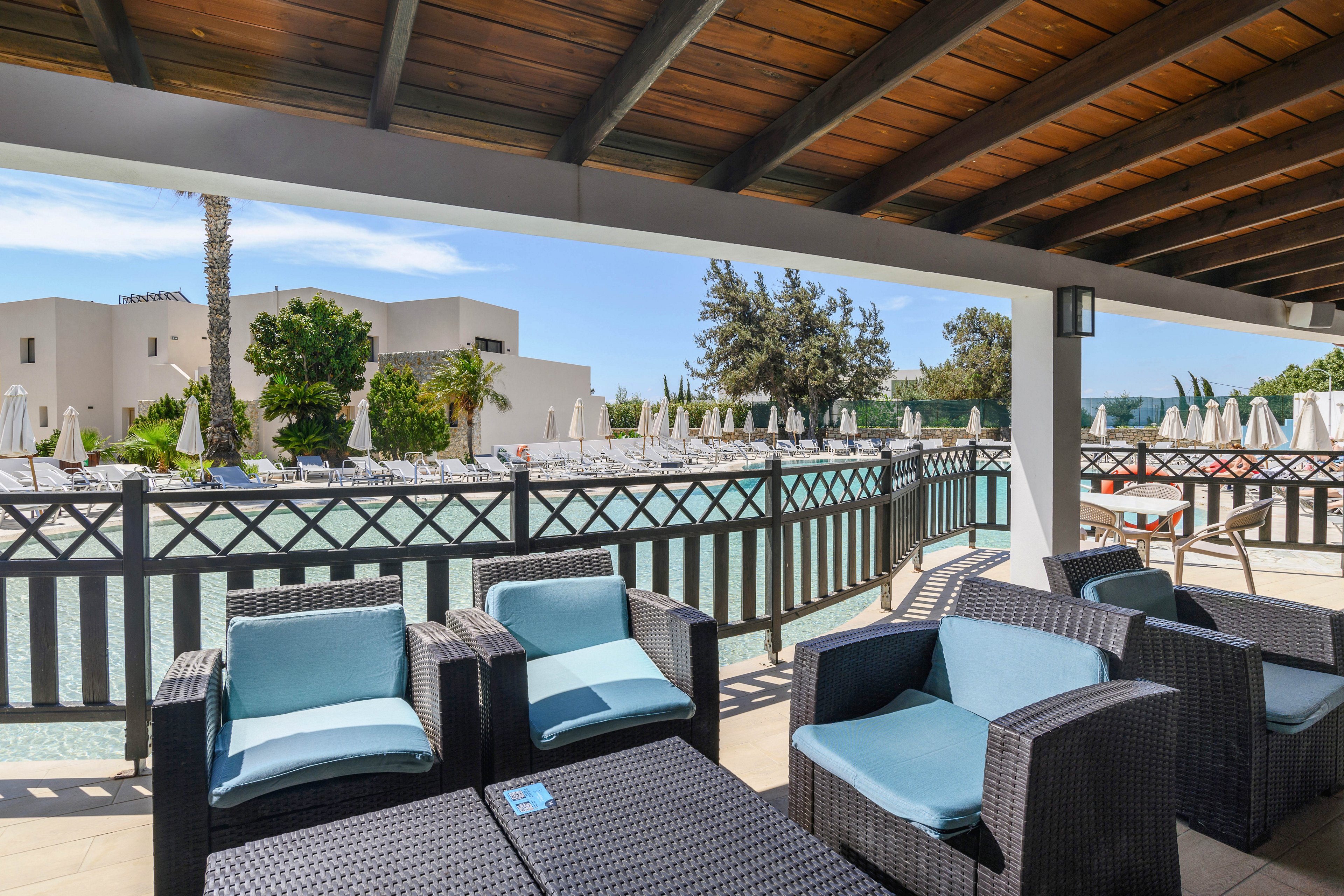 An outdoor lounge area with wicker armchairs and cushioned seats sits under a shaded wooden pergola. Beyond the railing, a large swimming pool is lined with white sun loungers and umbrellas. Modern white resort buildings and tall trees frame the background under a bright blue sky. The scene suggests a relaxing vacation setting with comfortable seating and calm water views.