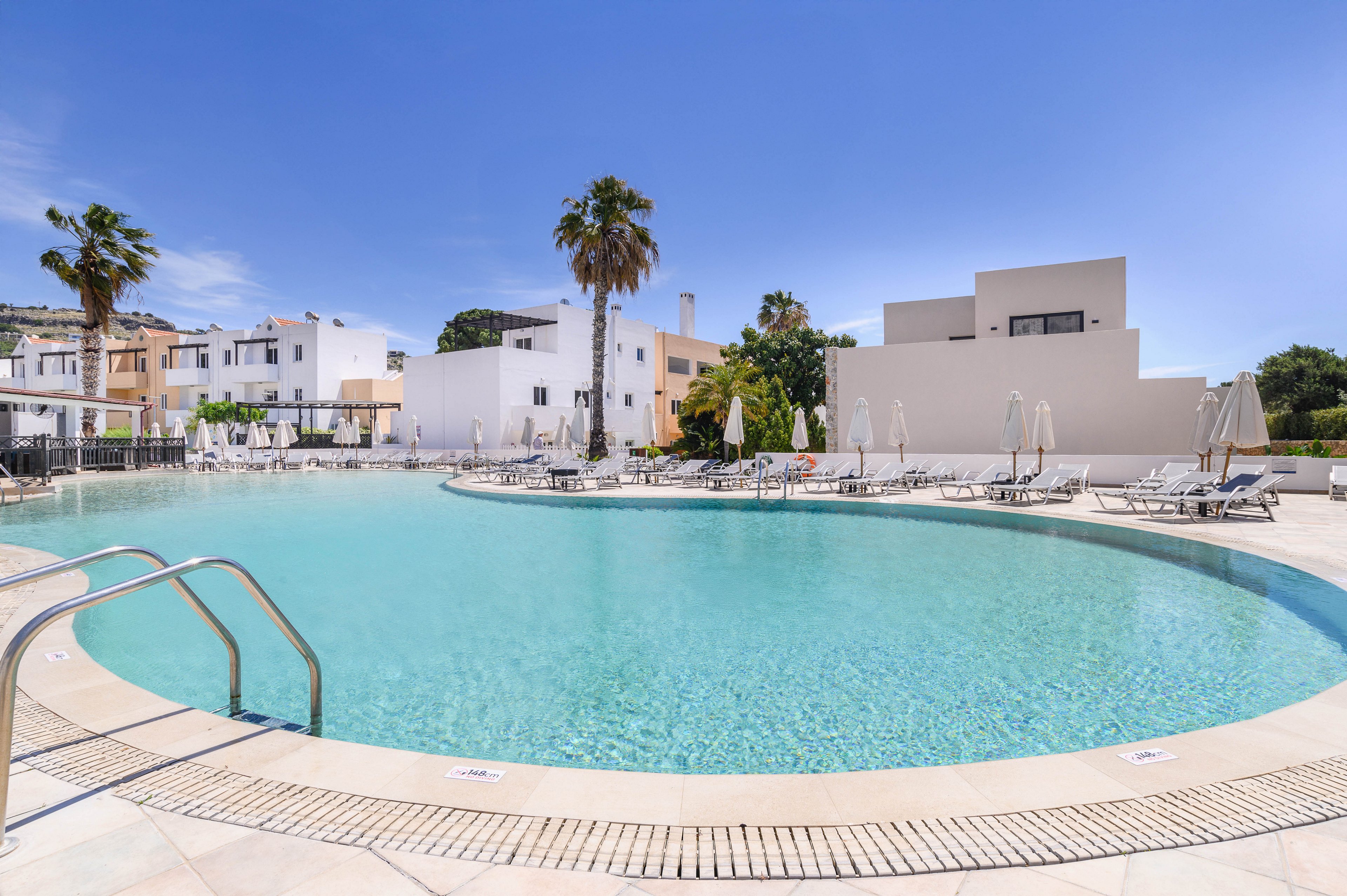 A large circular swimming pool sits at the center of a modern resort complex. White buildings and palm trees surround the pool under a clear blue sky. Numerous sun loungers and umbrellas line the poolside, suggesting a relaxing vacation setting. The scene is bright and tranquil, with no people visible in the area.