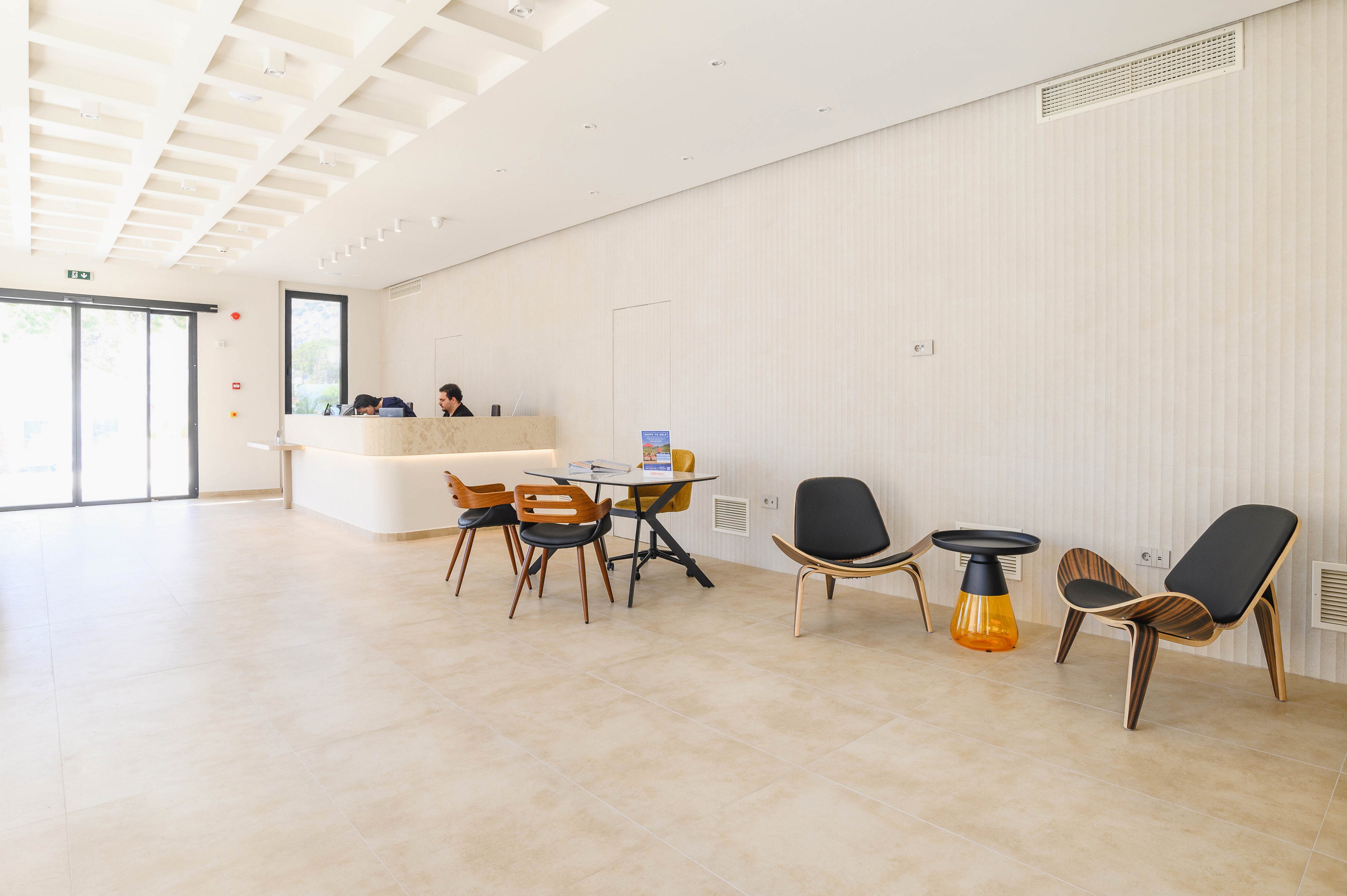 A bright, minimalist reception area features a curved white front desk with a person working behind it. Modern wooden and black chairs are arranged around small side tables in the open space. Large floor-to-ceiling windows on the left allow natural light to flood the room. The neutral color palette and clean lines create a calm, contemporary atmosphere suitable for an office or hospitality setting.
