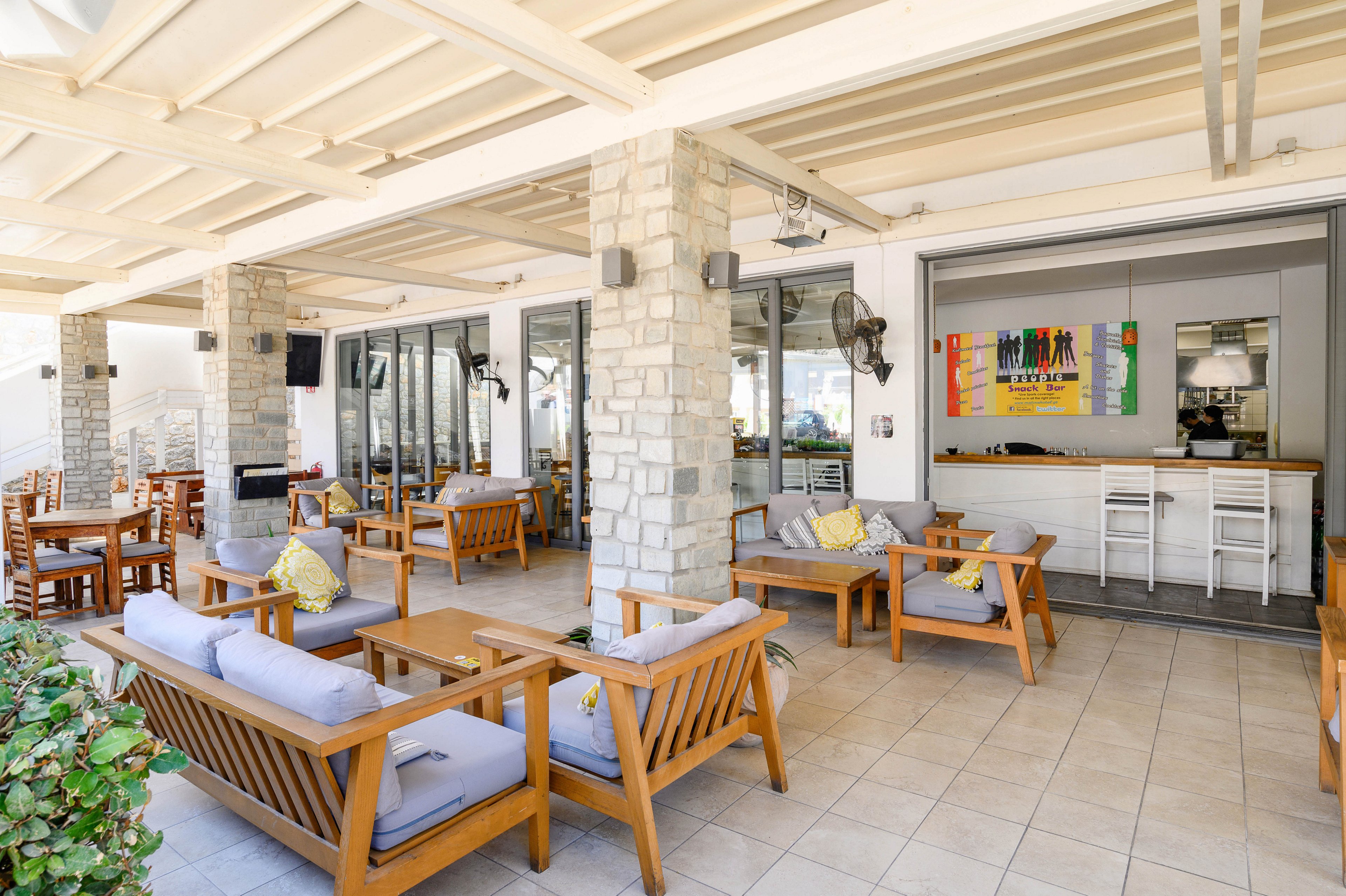 An open-air cafe lounge features wooden sofas with gray cushions arranged around low tables. The space is covered by a light-colored pergola roof and supported by stone columns. Large glass doors lead to an indoor bar area with high stools and colorful wall art. The setting appears calm and inviting, suitable for casual dining or relaxing with drinks.