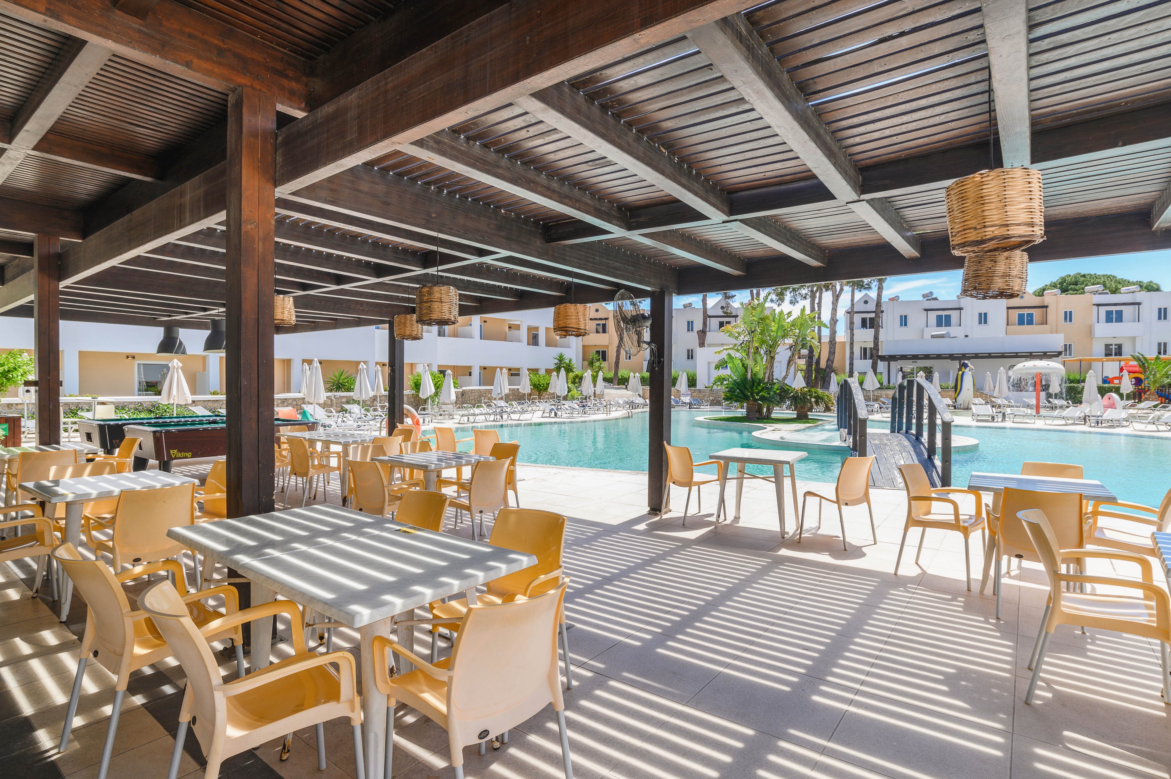 An open-air dining terrace with yellow chairs and white tables overlooks a large swimming pool. The space is shaded by a wooden pergola that casts striped shadows on the floor. Sun loungers and umbrellas line the poolside, with a small bridge crossing the water. In the background, low-rise resort buildings and palm trees create a relaxed holiday atmosphere under a clear sky.