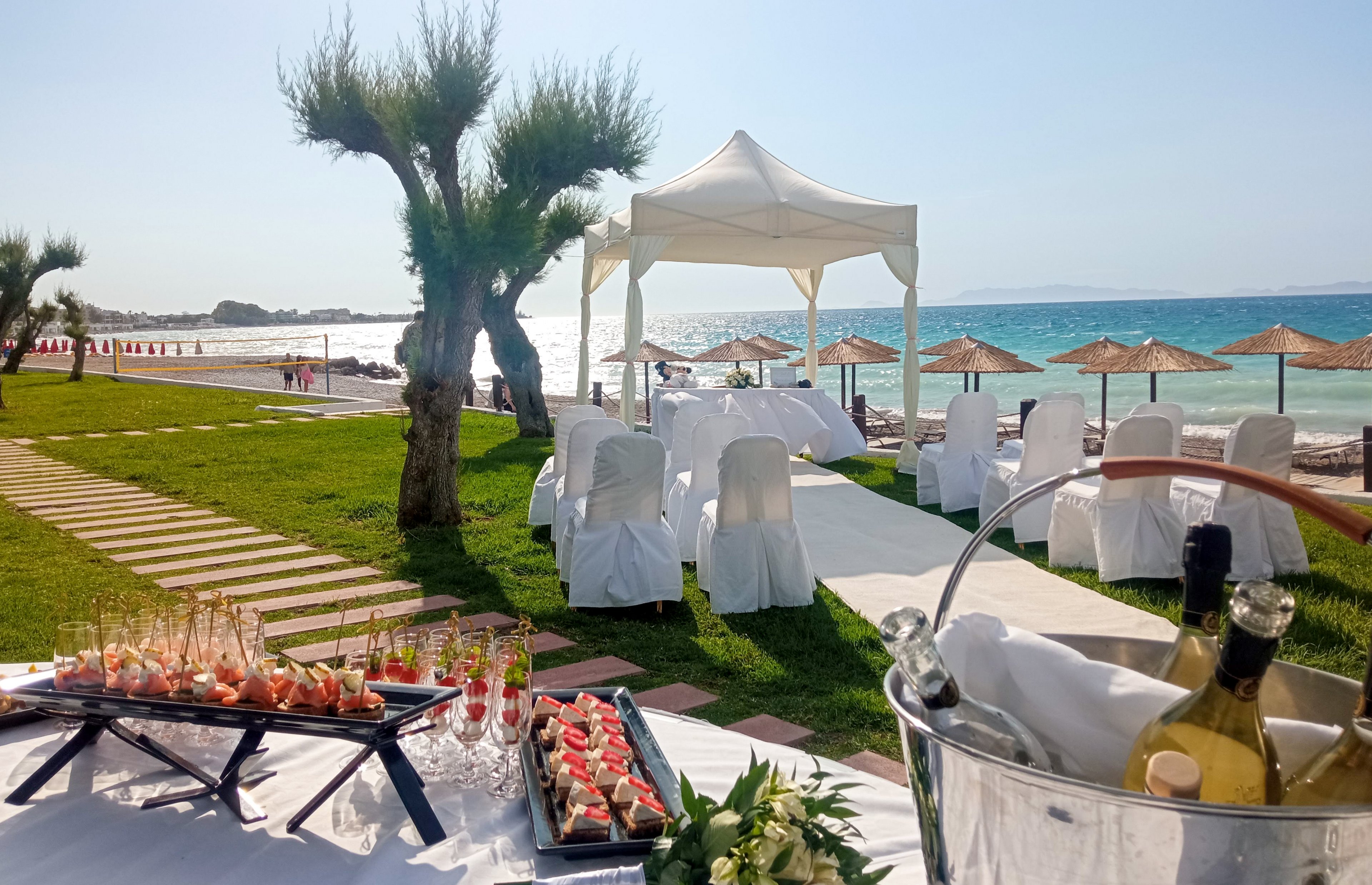An elegant wedding reception is arranged on a grassy lawn beside a bright blue sea. White-covered chairs and tables sit beneath a canopy facing the shoreline, with straw beach umbrellas visible in the background. In the foreground, trays of appetizers and rows of champagne flutes are prepared for guests. A metal ice bucket holds chilled bottles of white wine, enhancing the festive atmosphere under clear sunny skies.
