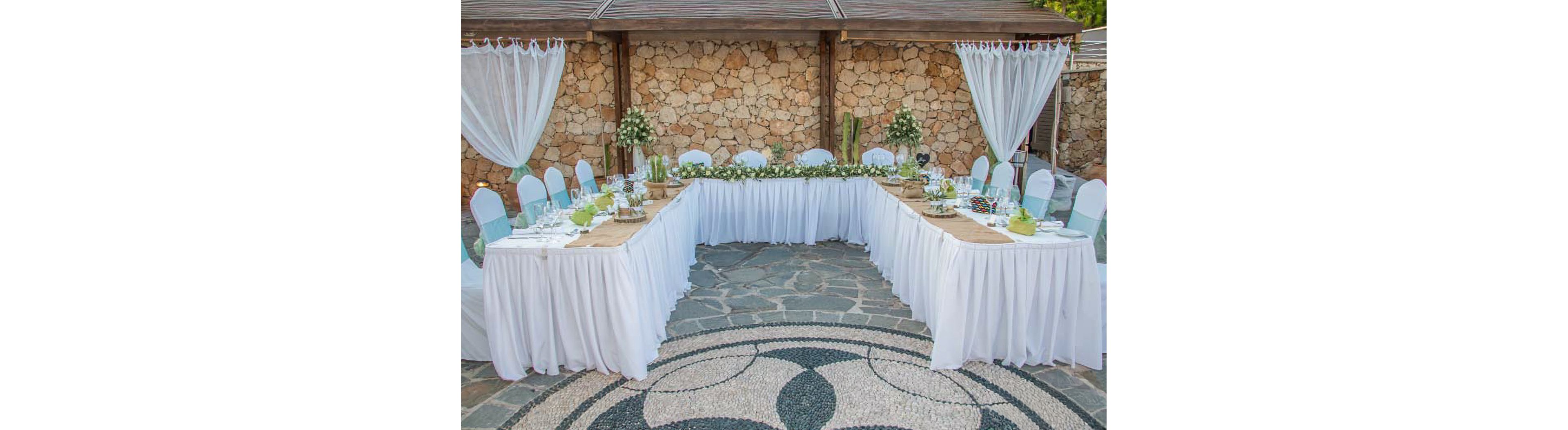 An elegant U-shaped table arrangement is set for an outdoor event against a rustic stone wall backdrop. White tablecloths and chair covers with light blue sashes create a clean, sophisticated look. The table is decorated with greenery, white floral centerpieces, and neatly arranged place settings. Sheer white curtains and potted plants enhance the romantic, intimate atmosphere of the venue.