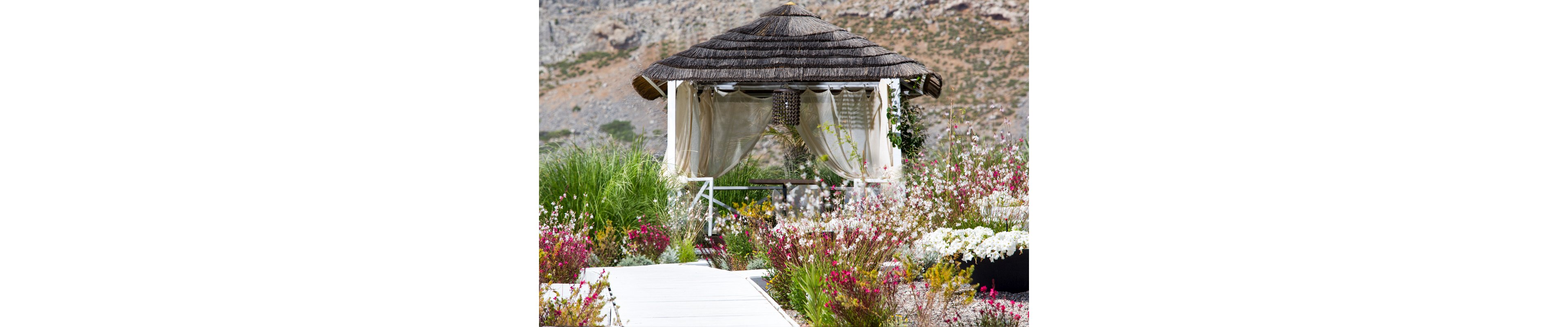 A thatched-roof gazebo with light, flowing curtains stands in the middle of a lush garden. A white pathway leads toward the structure, surrounded by colorful blooming flowers and tall ornamental grasses. The gazebo appears inviting and shaded, offering a tranquil spot to rest. Rocky hills rise softly in the background, enhancing the serene outdoor atmosphere.