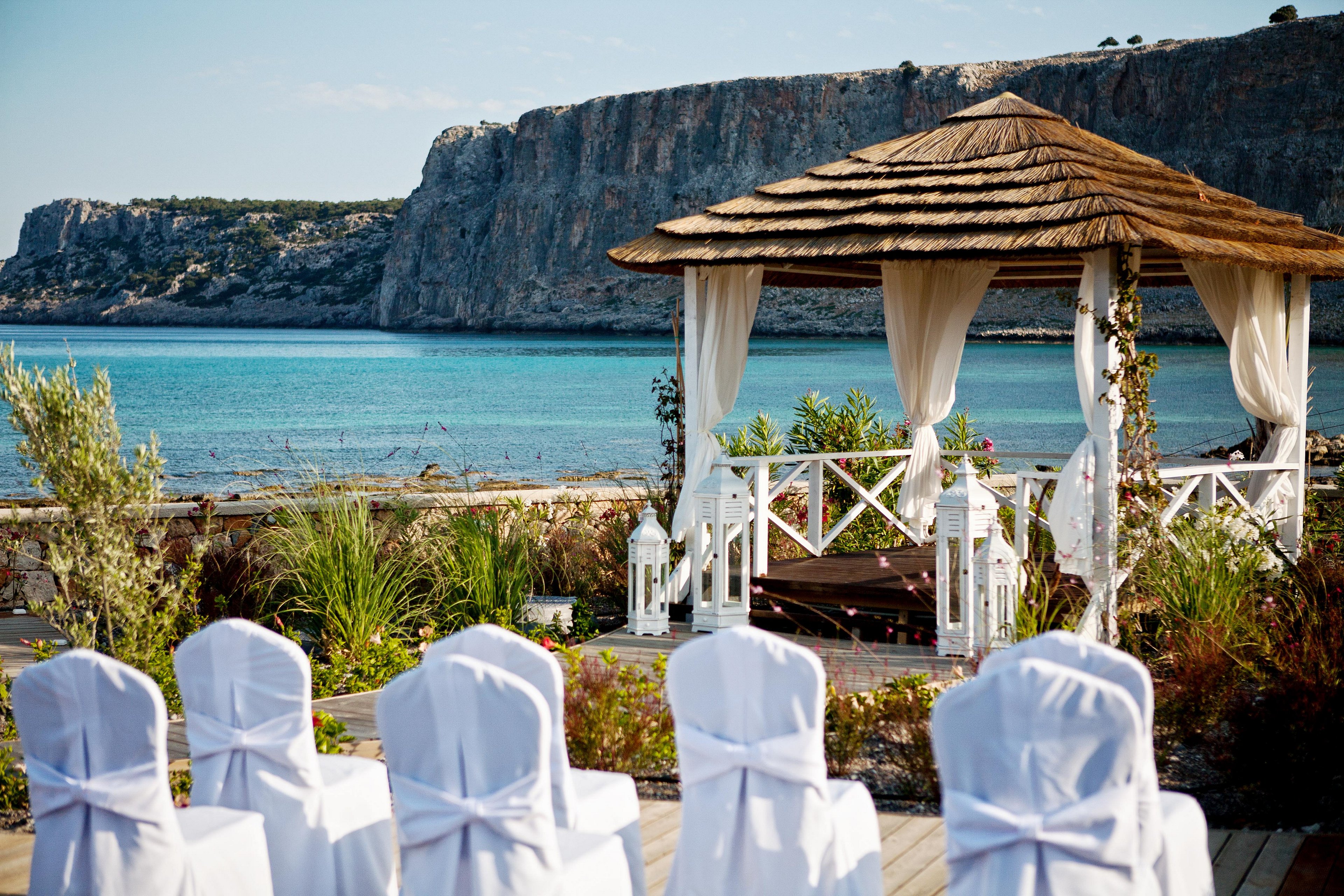 A wooden gazebo draped with white curtains stands beside a calm turquoise sea. Rows of white-covered chairs face the gazebo, suggesting a wedding ceremony setup. Lush green plants and lanterns decorate the foreground, adding a romantic touch. Rocky cliffs and a clear blue sky create a dramatic coastal backdrop.