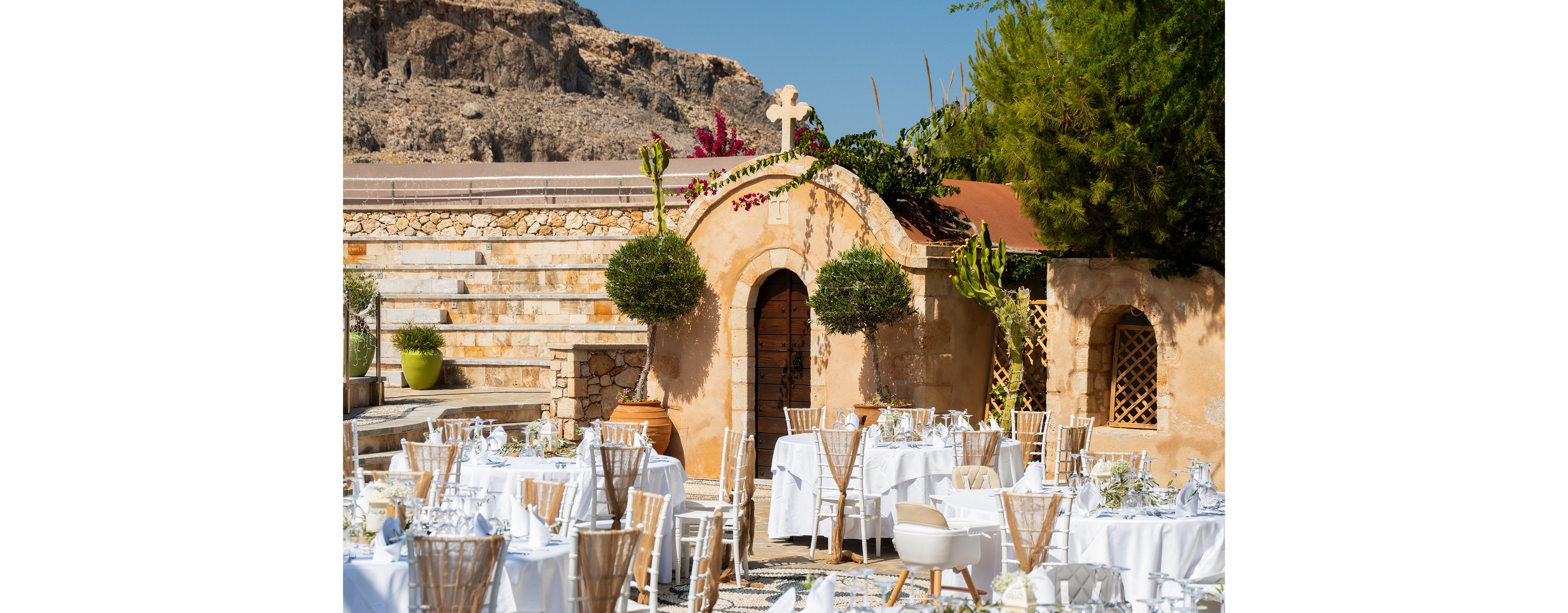 An elegant outdoor wedding reception is set up in front of a small rustic chapel with stone walls and a prominent cross above the entrance. Round tables covered in white linens are arranged with glassware, plates, and floral centerpieces, surrounded by wooden chairs. Lush greenery, potted plants, and manicured trees frame the courtyard, contrasting with the warm earthy tones of the building. Rocky hills rise in the background under bright, clear daylight, creating a picturesque Mediterranean atmosphere.