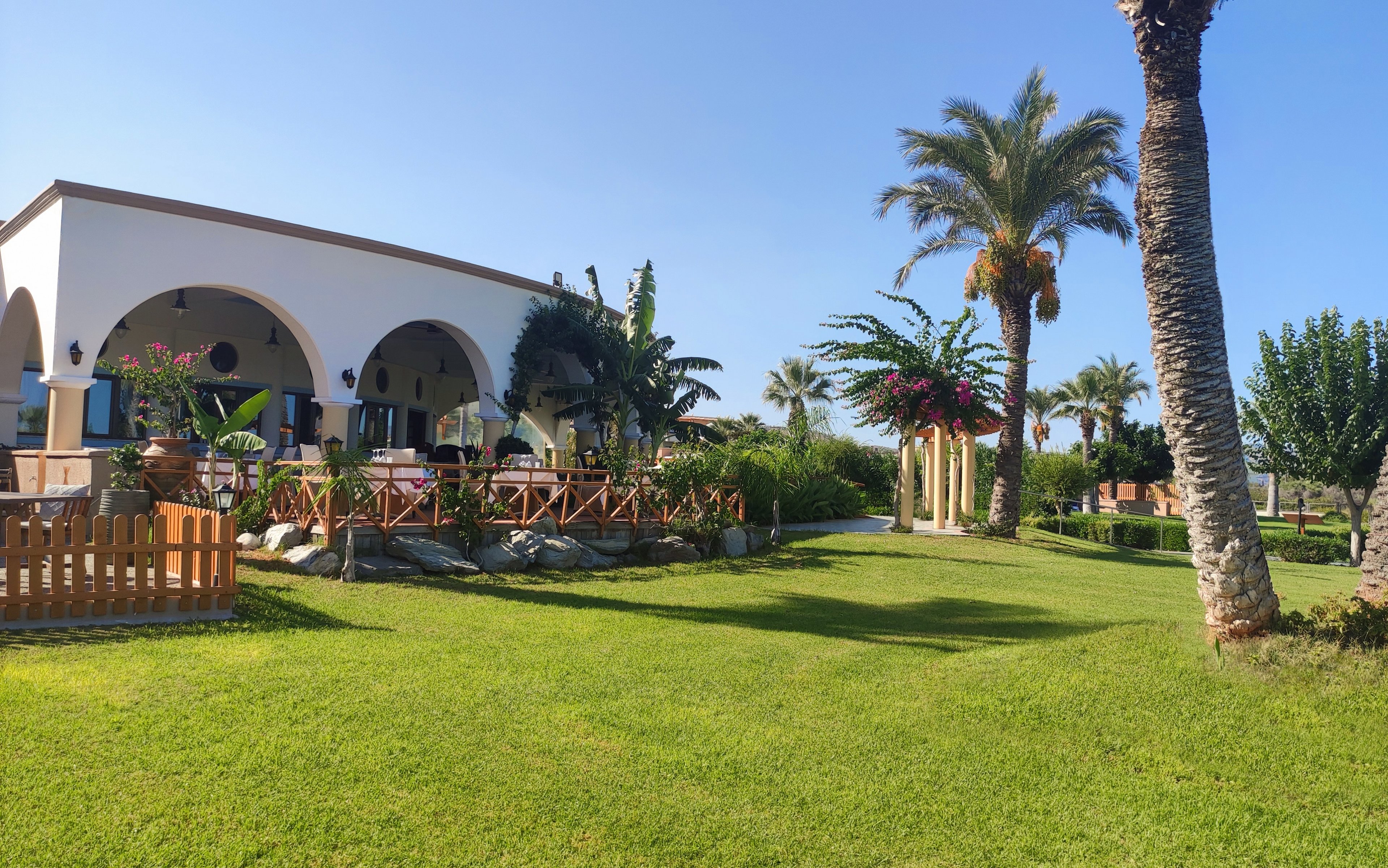 A white villa with large arched terraces stands beside a well-kept lawn under a clear blue sky. Palm trees and flowering shrubs surround the building, creating a tropical resort atmosphere. Wooden fences and stone borders define the garden areas and pathways. The bright sunlight and vivid greenery suggest a warm, relaxing holiday setting.