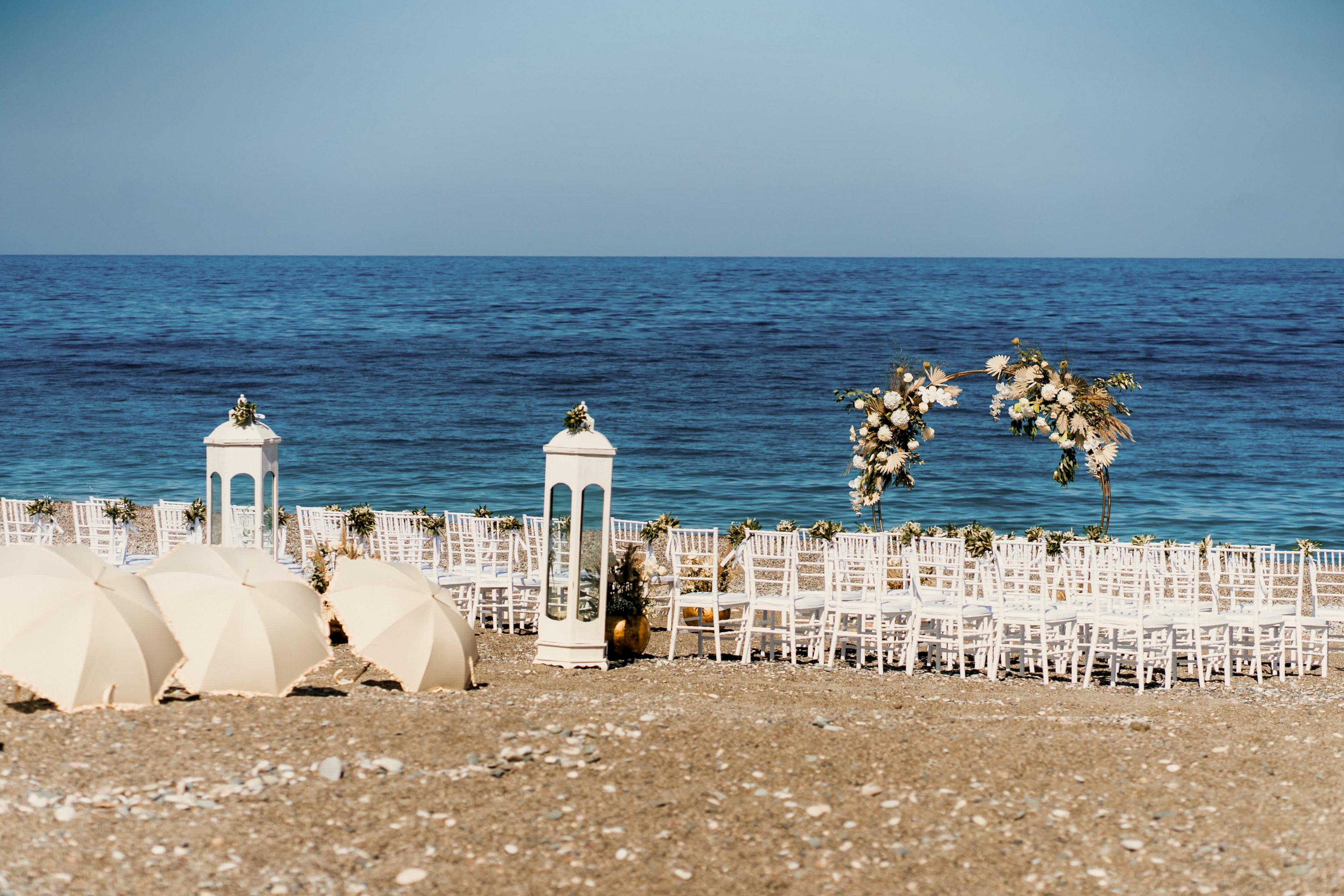 A beautifully arranged wedding ceremony is set up on a rocky beach facing the calm blue sea. White chairs are lined up in rows, leading to a floral arch decorated with white and blush flowers and greenery. Large white lanterns stand beside the seating area, adding a romantic decorative touch. Closed white umbrellas rest on the pebbled shore, and the clear sky enhances the serene coastal atmosphere.