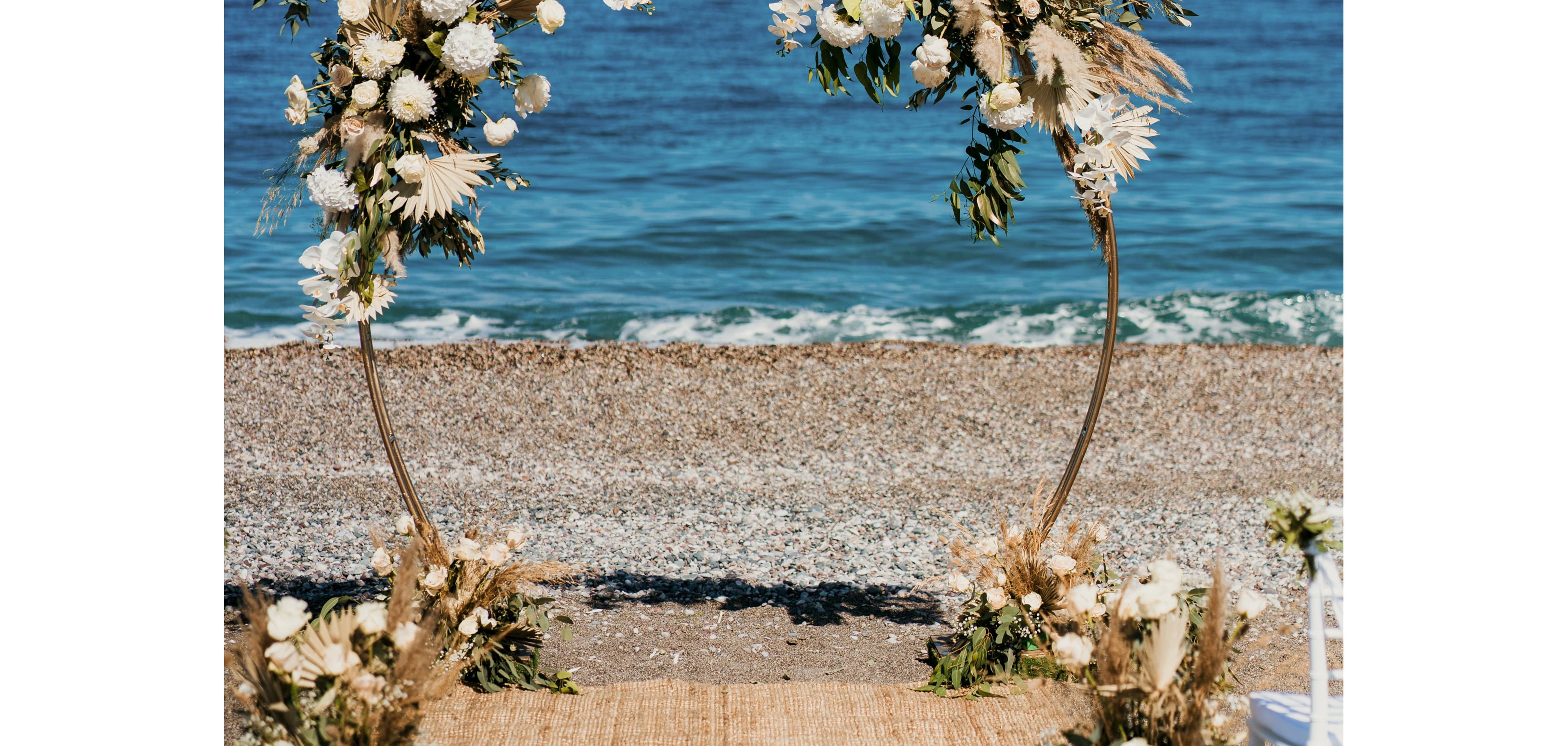 A circular wedding arch decorated with white flowers and dried foliage stands on a sandy beach. The arch frames the shoreline where gentle waves meet the pebbled coast. A woven aisle runner leads toward the arch, suggesting a ceremony setup. The bright blue sea and clear sky create a serene, romantic atmosphere.