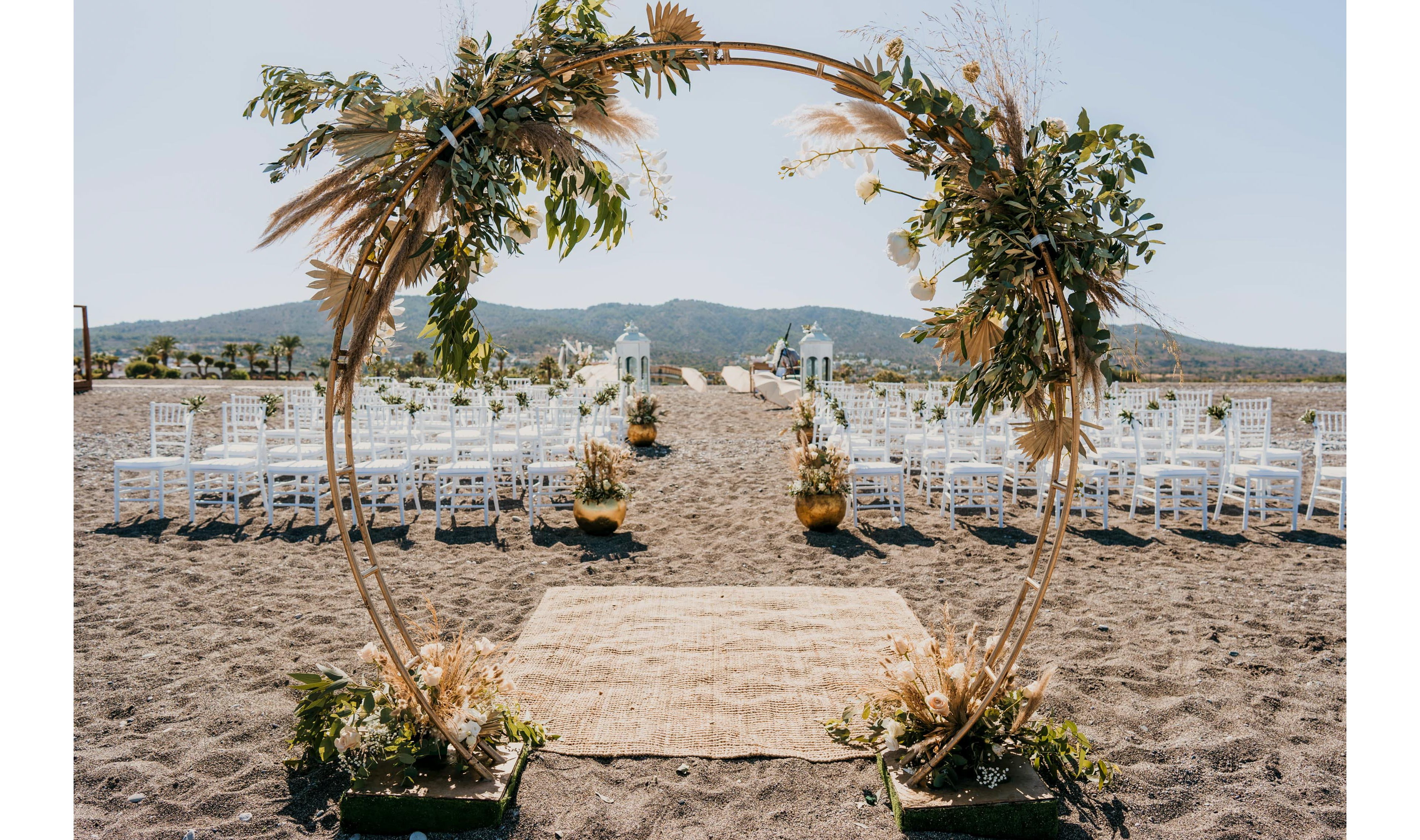 A circular floral arch stands on a sandy beach, framing the aisle for an outdoor wedding ceremony. White chairs are arranged in neat rows on either side of a natural fiber runner leading toward the water. Dried grasses, greenery, and potted plants decorate the arch and aisle, creating a rustic boho style. Distant hills and a clear blue sky complete the serene coastal backdrop.