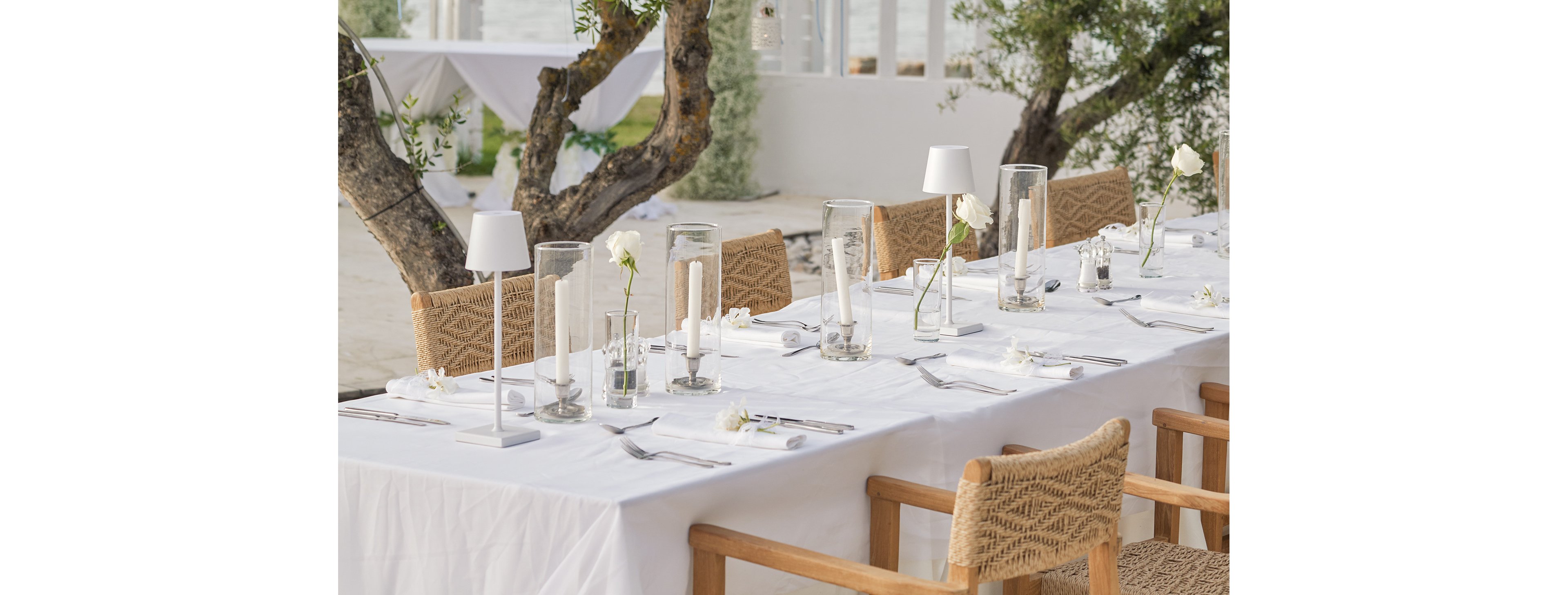 A long white-clothed table is elegantly set for an outdoor gathering. Glass vases with white flowers and tall candles create a minimalist centerpiece arrangement. Woven chairs surround the table, and small white lamps add a modern touch. The setting appears to be near the water, with trees and soft natural light enhancing the serene atmosphere.