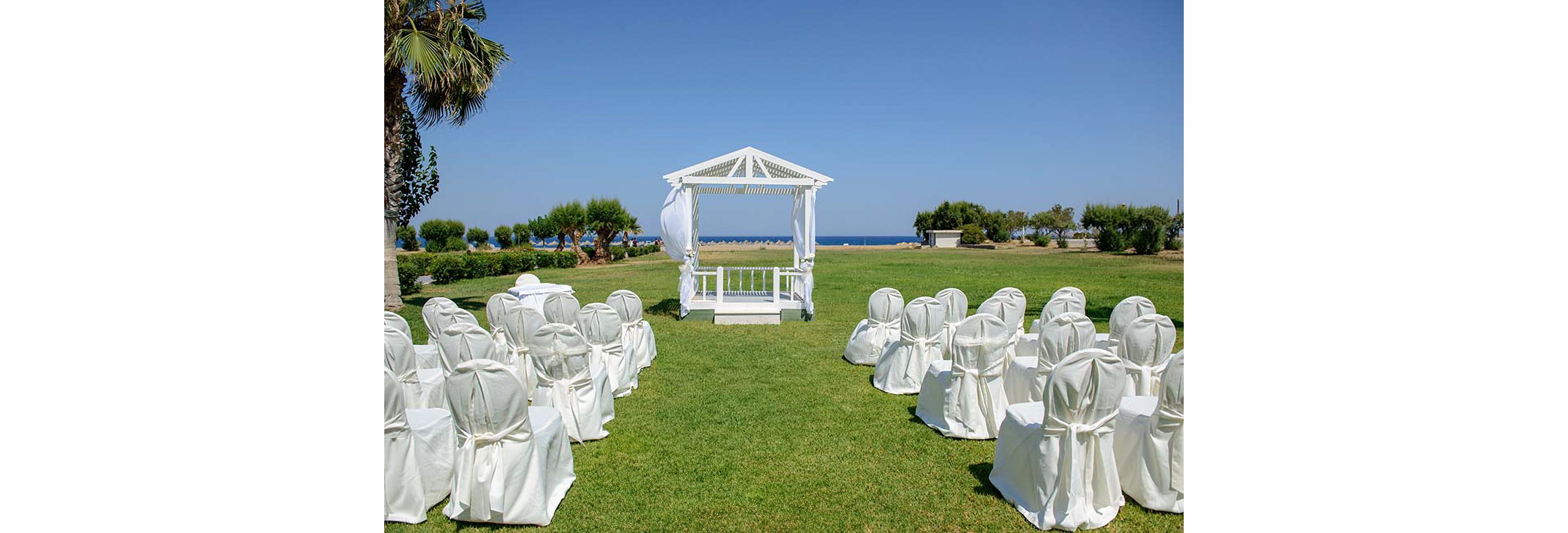 An outdoor wedding ceremony area is arranged on a green lawn facing the sea. A white gazebo stands at the center, draped with light fabric and framed by a clear blue sky. Rows of white chair covers are neatly aligned on both sides, creating an aisle leading to the gazebo. Trees and shrubs border the lawn, with the ocean visible in the background.