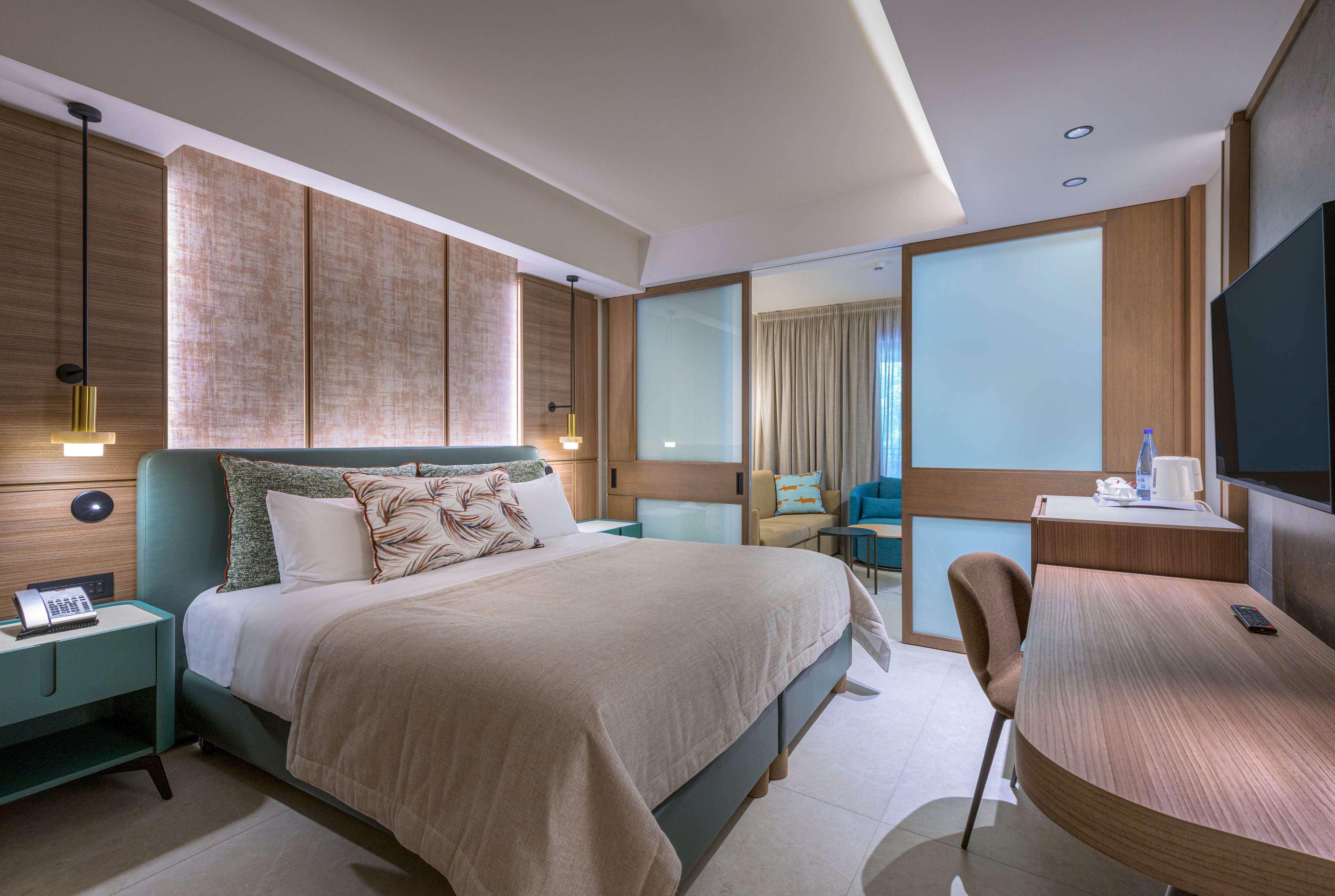A contemporary hotel bedroom features a large bed with neutral bedding and decorative pillows. The room includes a wooden desk with a chair, bedside tables, and pendant lights that create a warm ambiance. Sliding glass doors reveal a separate sitting area with a sofa and armchair, softly lit by natural light through curtains. A wall-mounted television and minimalist decor emphasize comfort and modern style.