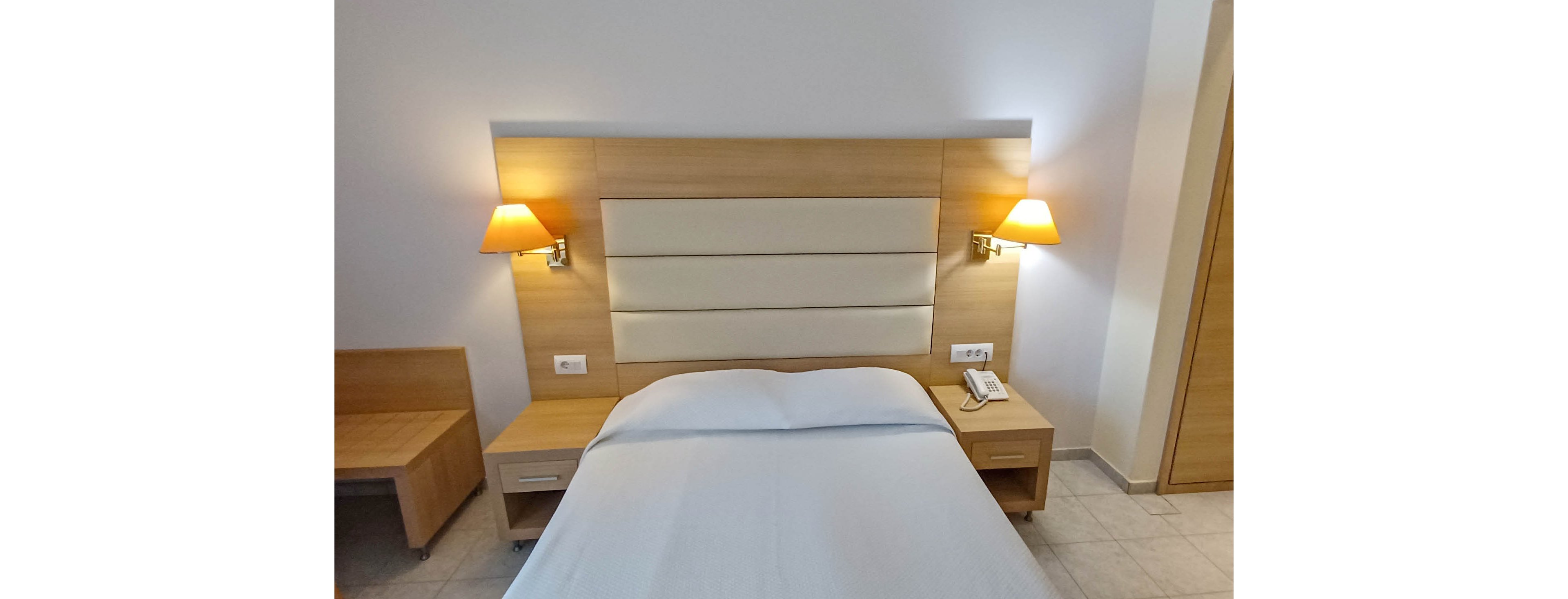A neatly made single bed is centered against a light wood headboard in a compact hotel room. Two warm wall-mounted lamps flank the bed, casting a soft glow on the neutral walls. A bedside table with a telephone and power outlets sits to one side, while a simple chair and desk occupy the corner. Light-colored flooring and minimal decor create a clean, modern, and functional atmosphere.
