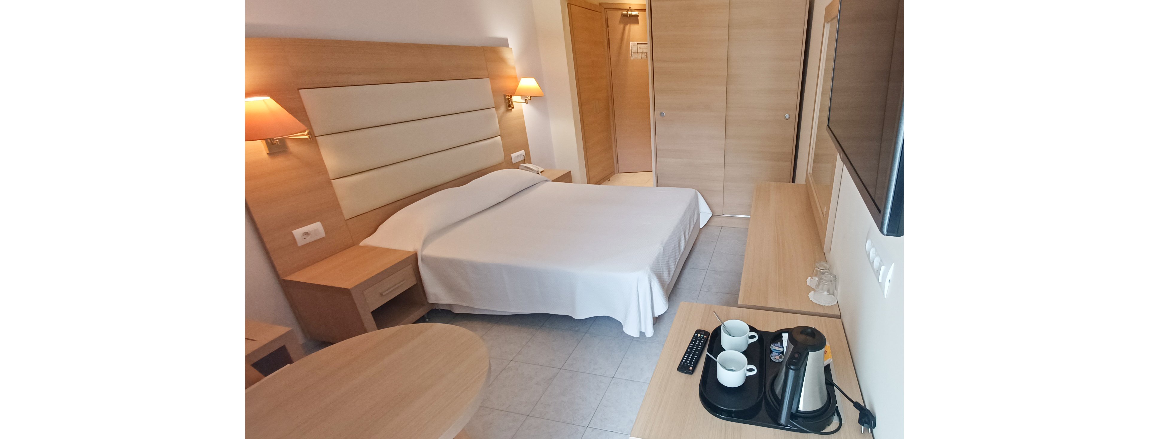 A neatly made double bed is the focal point of this modern hotel room. Warm wall lamps and light wood furniture create a comfortable, inviting atmosphere. A flat-screen television is mounted on the wall opposite the bed, above a long desk. On the desk, a tray holds a kettle, cups, and complimentary beverages, emphasizing guest convenience.