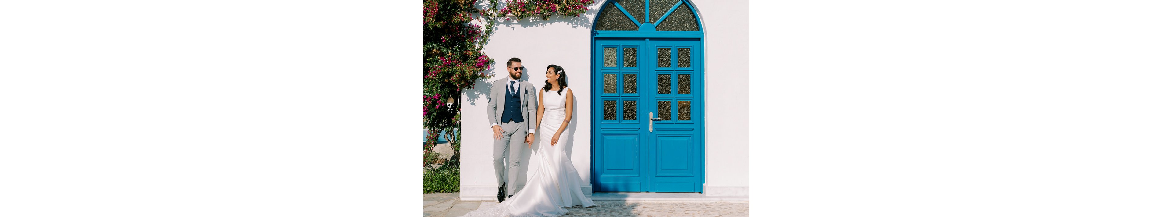A bride in a fitted white wedding gown stands beside a groom in a light gray suit and navy vest. They pose casually against a white wall next to a large bright blue arched door. Sunlight creates crisp shadows on the ground, highlighting the clean architectural lines. Colorful flowering vines climb the wall, adding a romantic touch to the outdoor scene.