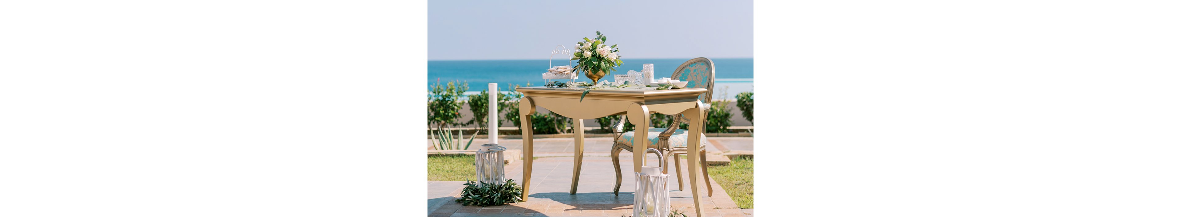 An ornate table with curved legs is set outdoors overlooking a bright blue sea. A matching upholstered chair with a floral pattern sits beside the table. The tabletop features white tableware, decorative cups, and a central bouquet of white and green flowers. Lanterns and greenery are arranged around the table, creating a refined coastal celebration atmosphere.