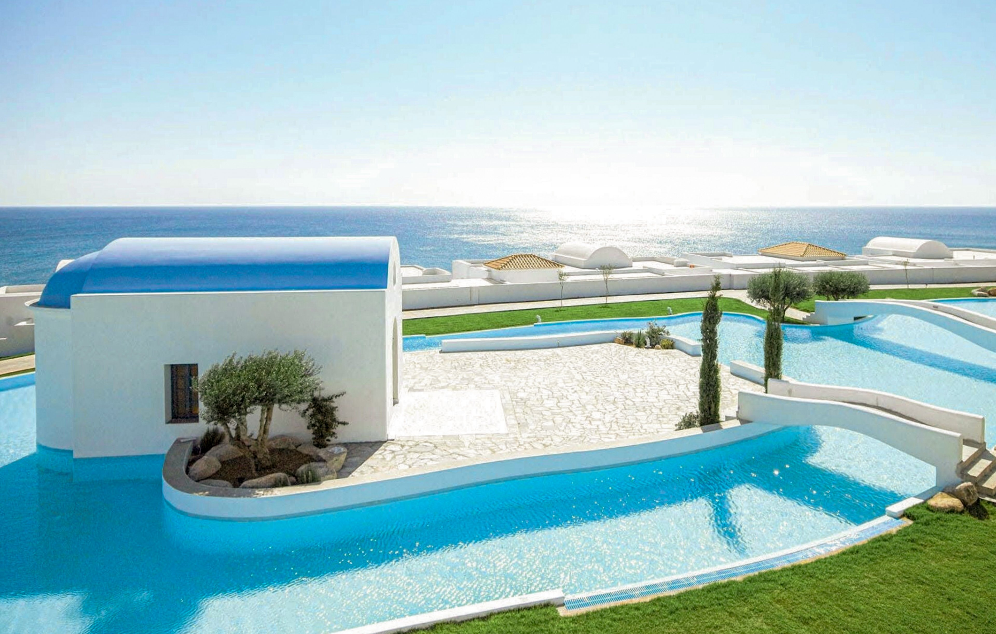 A modern white villa with a blue roof overlooks a calm, expansive sea. Curved infinity-style swimming pools surround the building, reflecting the bright sky. Minimalist landscaping with trimmed trees and green lawn contrasts with the white stone terrace. The scene suggests a luxurious, tranquil coastal resort under clear daylight.