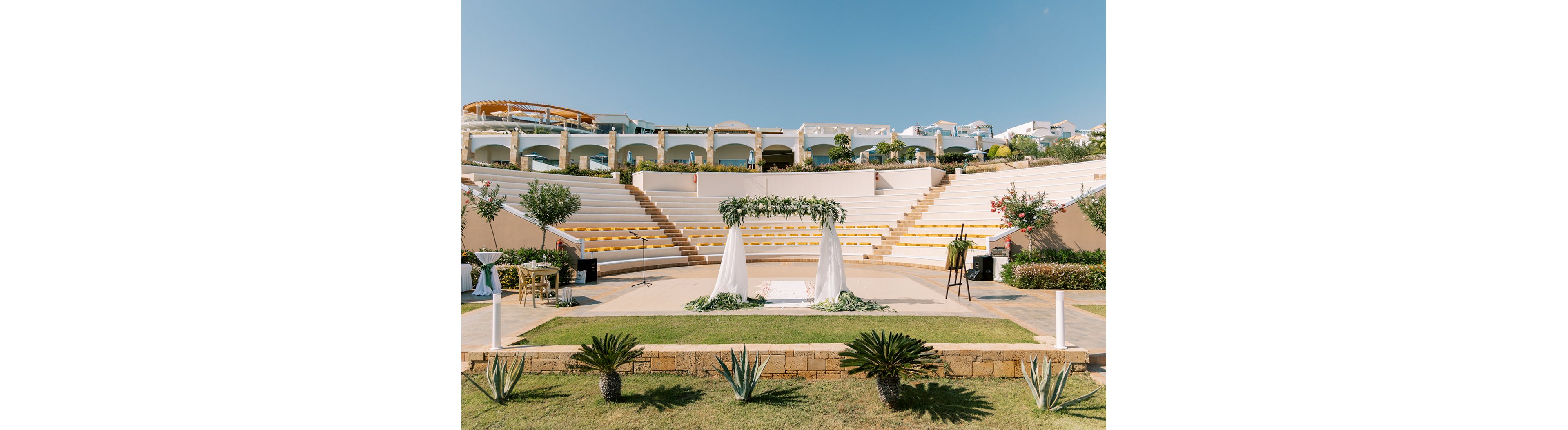 An outdoor amphitheater is prepared for a wedding ceremony under a clear blue sky. A floral arch with white drapery stands at the center of the stone stage, surrounded by steps with yellow seating. Green plants and small trees frame the scene, adding a natural touch to the architectural backdrop. The bright daylight and soft colors create a warm, romantic atmosphere.