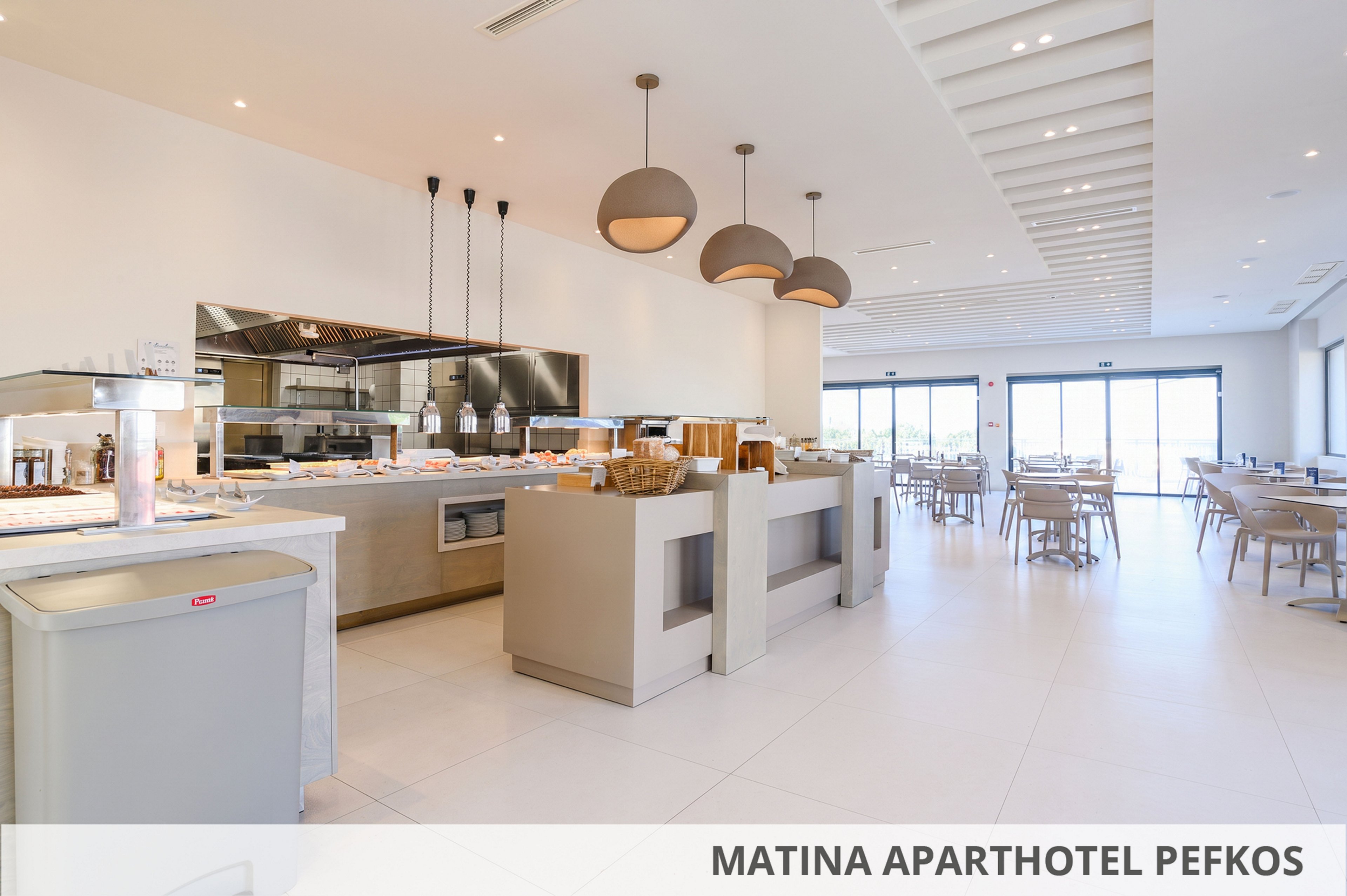 Modern self-service buffet area inside Matina Aparthotel Pefkos. The space features sleek counters, pendant lights, and an open kitchen with stainless steel equipment. Neutral tones and large windows create a bright, airy atmosphere with natural light. Several tables and chairs are arranged for guests in the adjoining dining area.