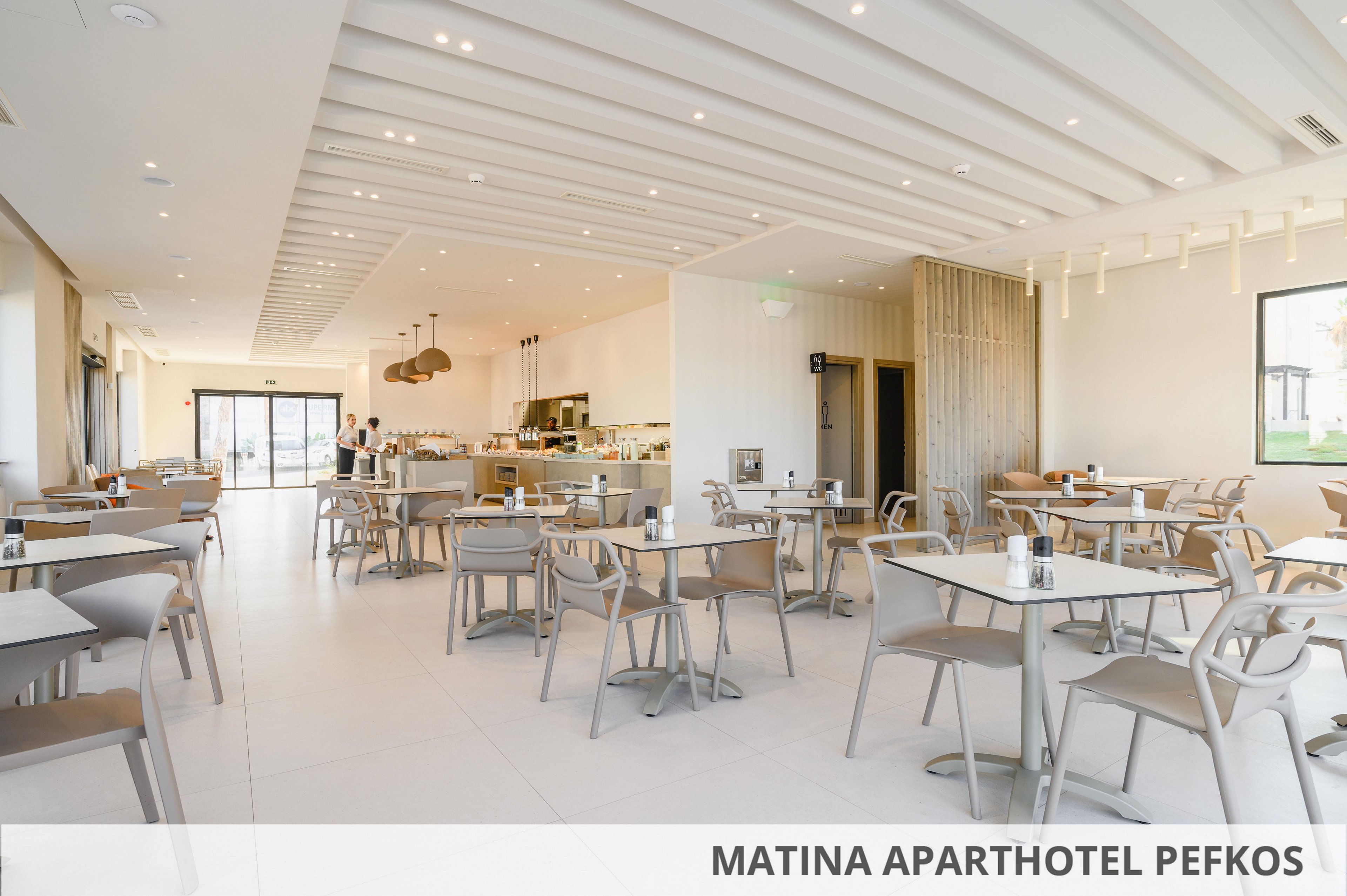 Spacious contemporary restaurant interior at Matina Aparthotel Pefkos with neatly arranged tables and chairs. The room features large windows, light-colored flooring, and a clean minimalist ceiling with recessed lighting. A buffet or bar counter is visible in the background, adding to the casual dining atmosphere. The overall setting feels airy, welcoming, and designed for relaxed hotel guests.